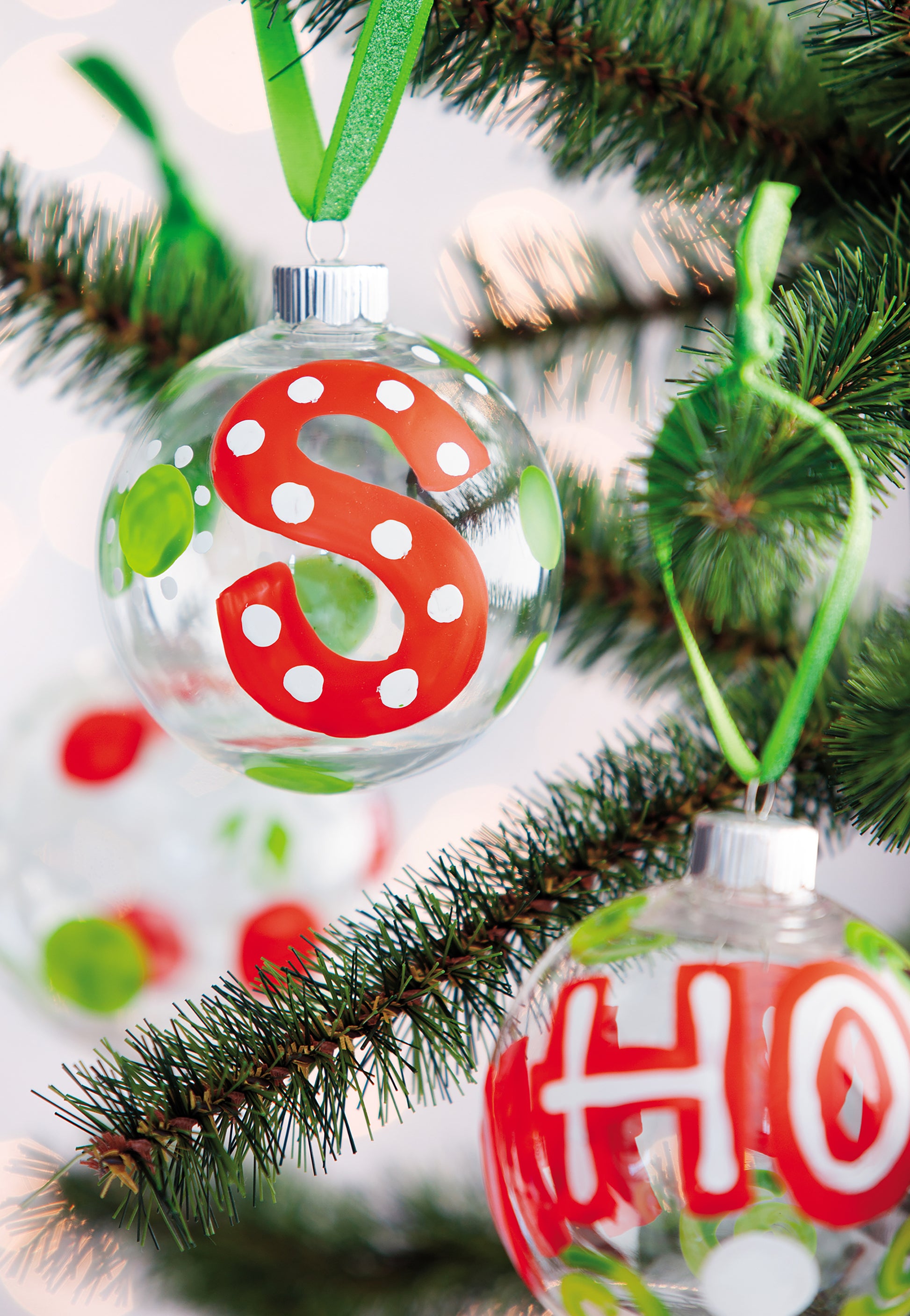 A close-up of ornaments crafted using the Holiday Ornaments Digital Download by Leisure Arts, featuring clear ornaments with red and white "S" and "HO" letters, green polka dots, ribbon loops, and pine needles in the background.