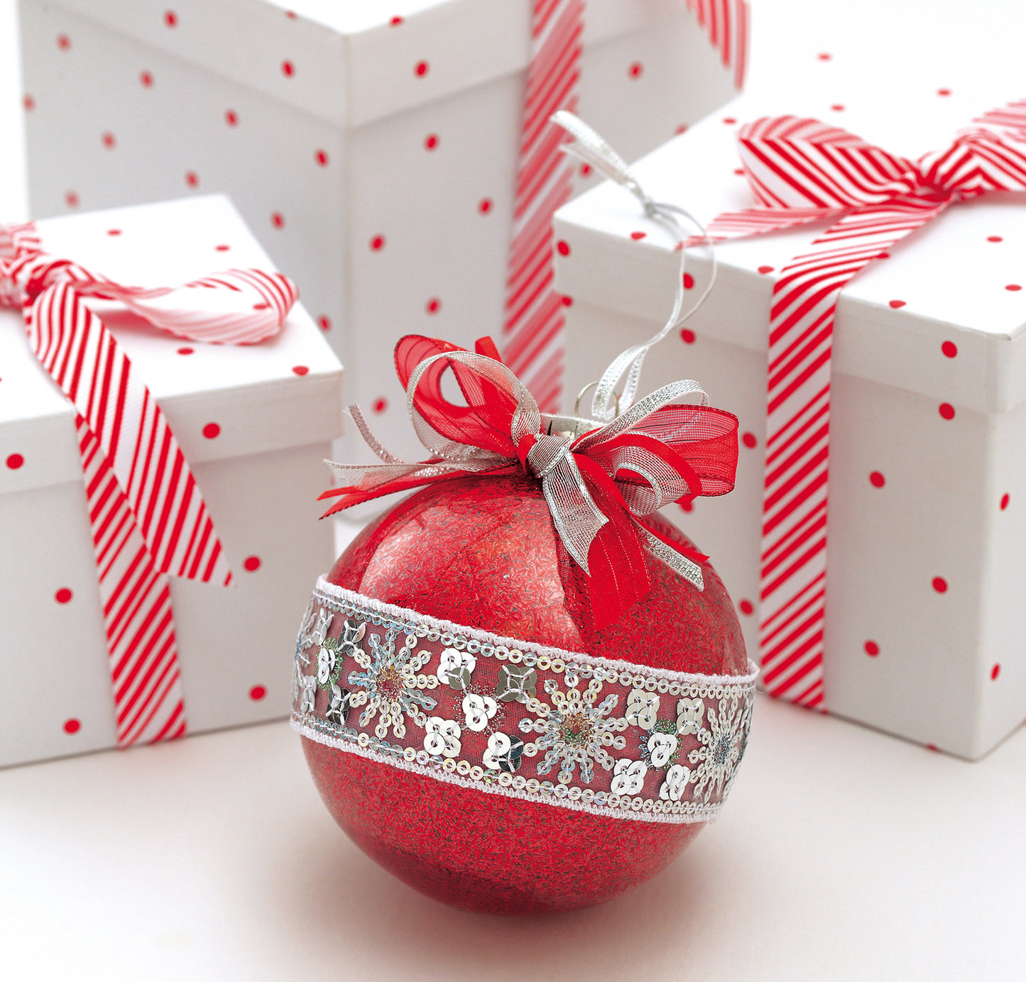 Get inspired for festive ornament crafts with the Holiday Ornaments Digital Download by Leisure Arts—featuring a red Christmas ornament with silver ribbon and lace, displayed before three white gift boxes adorned in holiday ribbons.