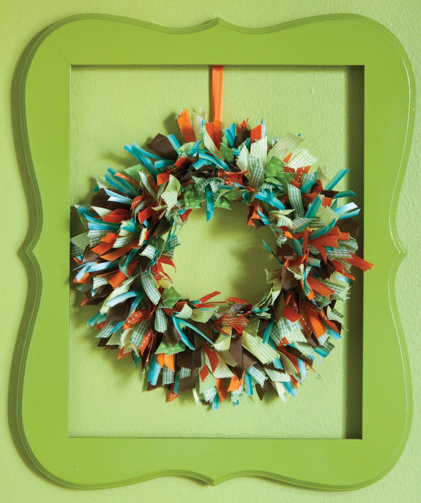 Add a unique touch to your home decor with the "Crafting with Buttons and Ribbons" kit by Leisure Arts—a colorful wreath of fabric strips in green, blue, orange, and brown framed in bright green on a light green wall.