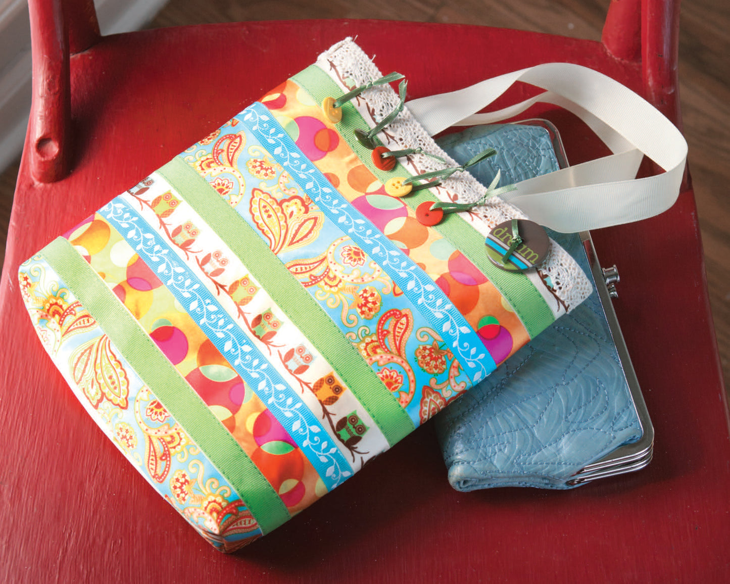 The Leisure Arts "Crafting with Buttons and Ribbons" tote bag features green, blue, orange, and yellow floral and paisley stripes with white handles—ideal for carrying needlework or paper crafts—next to a blue quilted wallet on a red chair.