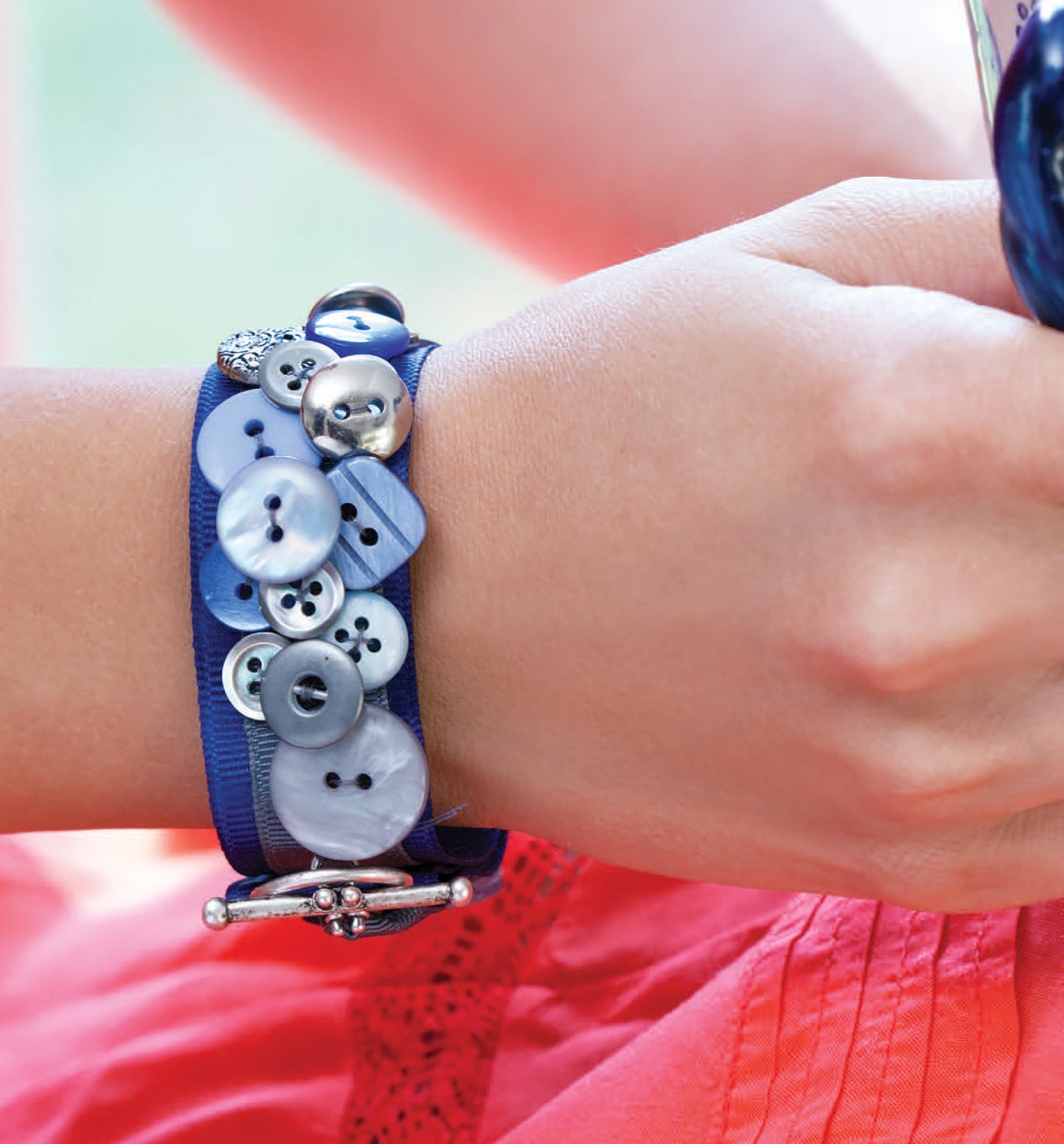 A wrist adorned with a blue, button-embellished bracelet appears beside a hand holding Crafting with Buttons and Ribbons by Leisure Arts, with a pink skirt visible—perfect inspiration for needlework or paper crafts.