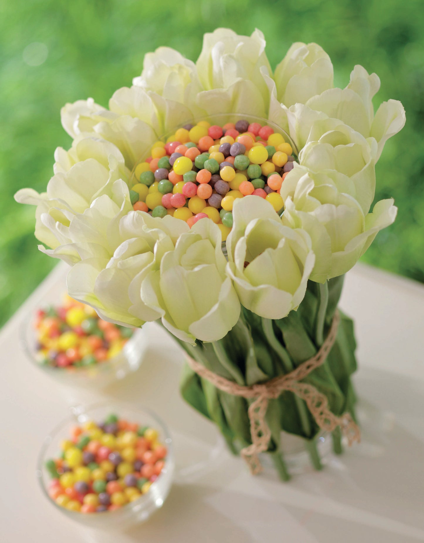 Create an easy DIY floral arrangement from "Crafting with Florals" by Leisure Arts—circle white tulips with colorful candies in the center for a cheerful home decor piece. Add two small glass bowls of candies on a white surface against a grassy backdrop.
