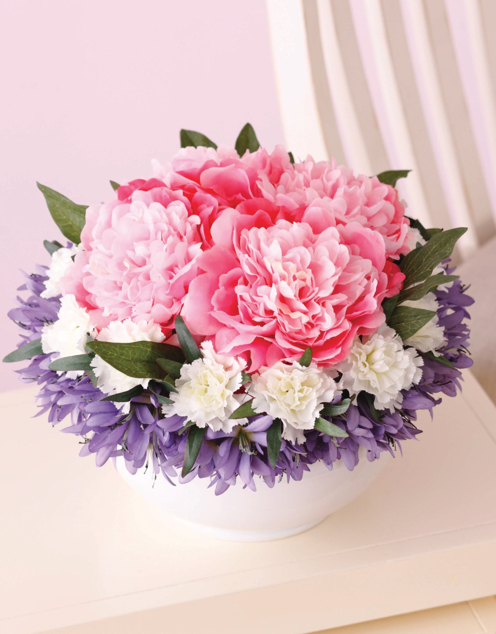 The "Crafting with Florals" arrangement by Leisure Arts features a white bowl filled with pink, white, and purple blooms, making it ideal for home decor or DIY enthusiasts. Displayed on a light surface with a white chair and soft pink background.