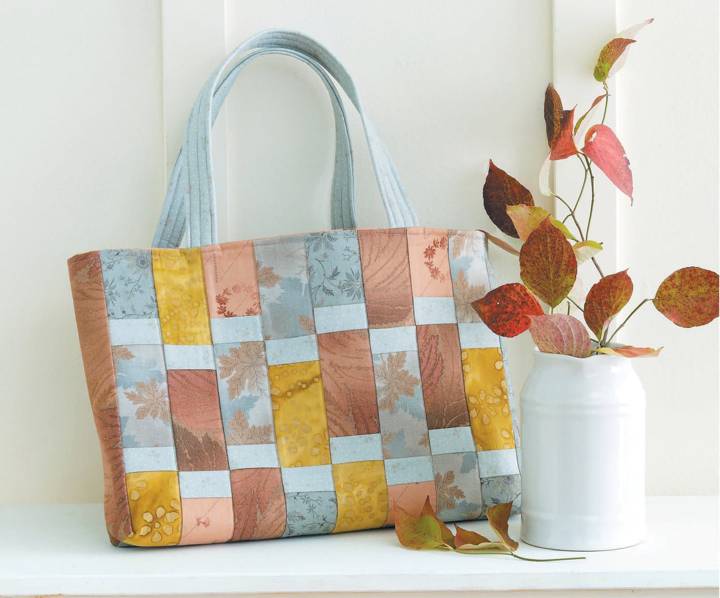 The Leisure Arts Fat Quarter Bags tote, with its patchwork design and pastel fabric inspiration, rests on a white shelf next to a white vase filled with reddish autumn branches.