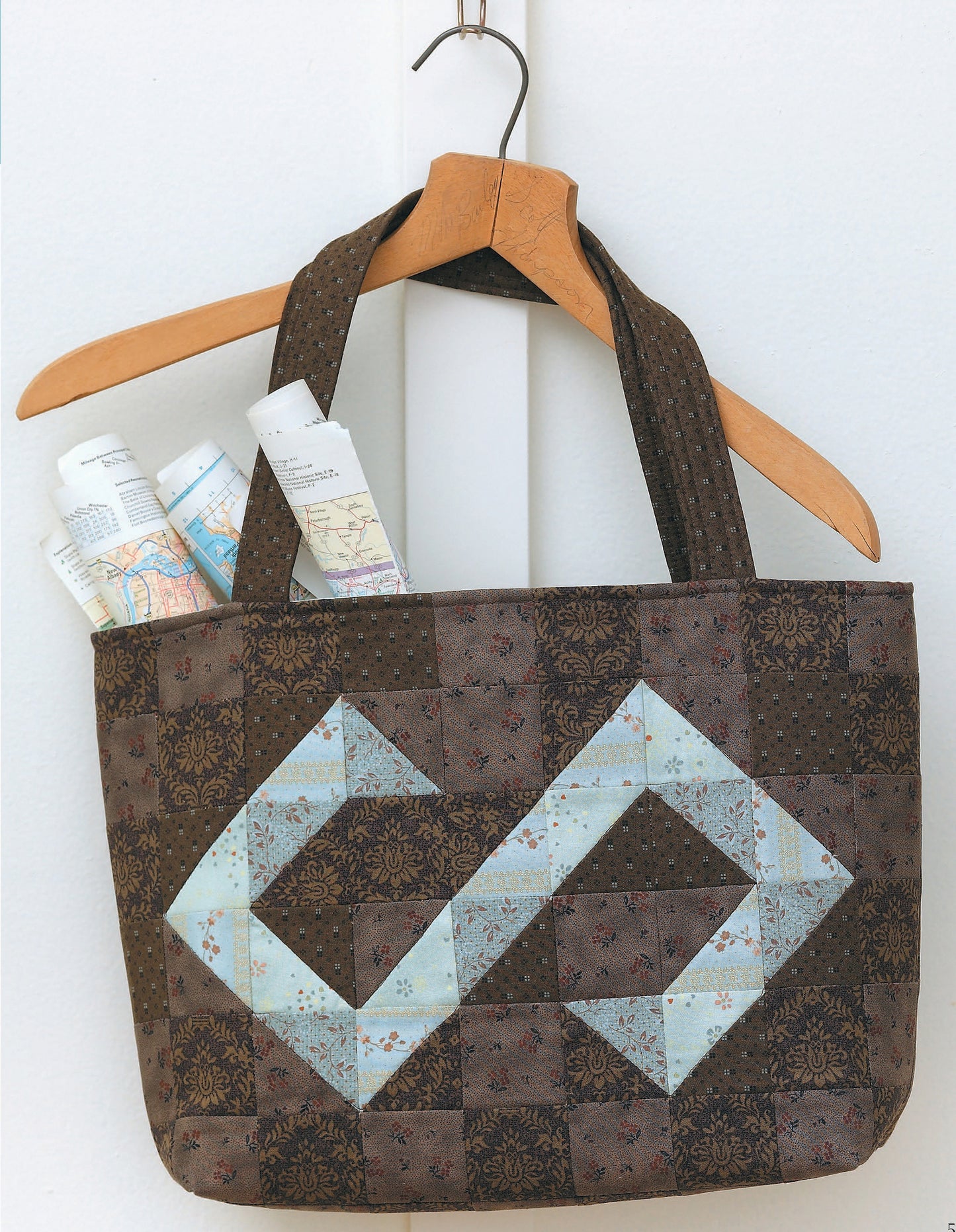 The Fat Quarter Bags by Leisure Arts feature a brown patchwork tote with light blue geometric designs, hanging on a wooden hanger. Its pocket holds maps and papers, perfect for fans of quilt-inspired fabric totes.
