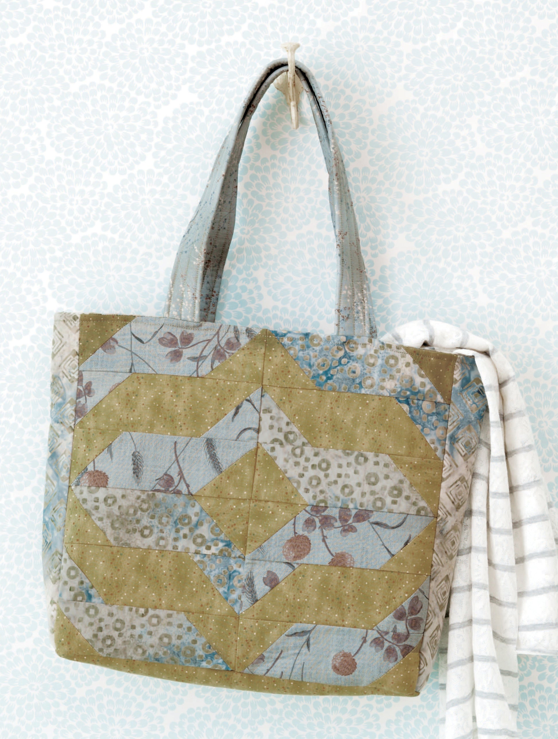 The Leisure Arts Fat Quarter Bags feature green and blue zigzag patterns, floral and polka dot prints, short handles, and a white-gray striped cloth tied on one handle—showcasing their charm on a hook against a pale patterned wall.