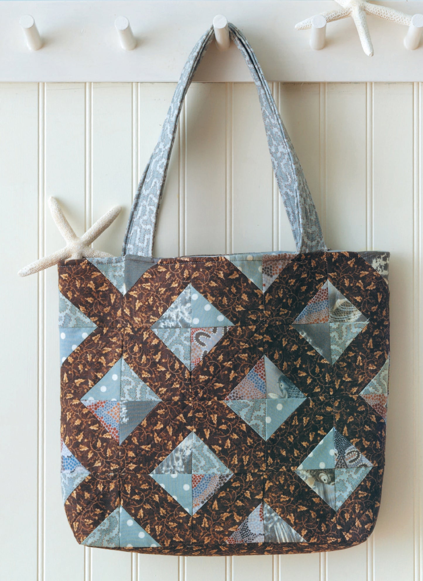 The Fat Quarter Bags by Leisure Arts, featuring geometric brown and blue patchwork, is ideal for fabric tote lovers. It hangs on a white peg rack adorned with starfish.