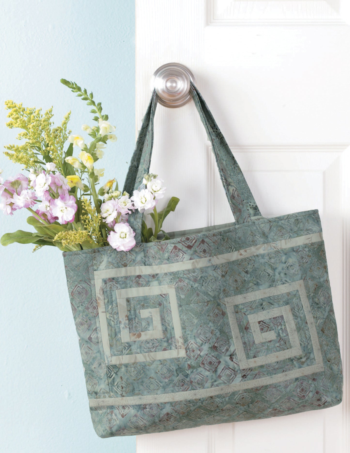 A Fat Quarter Bag from Leisure Arts with a green geometric pattern hangs on a doorknob, filled with yellow and purple flowers. The fresh white door and light blue wall highlight this charming tote.