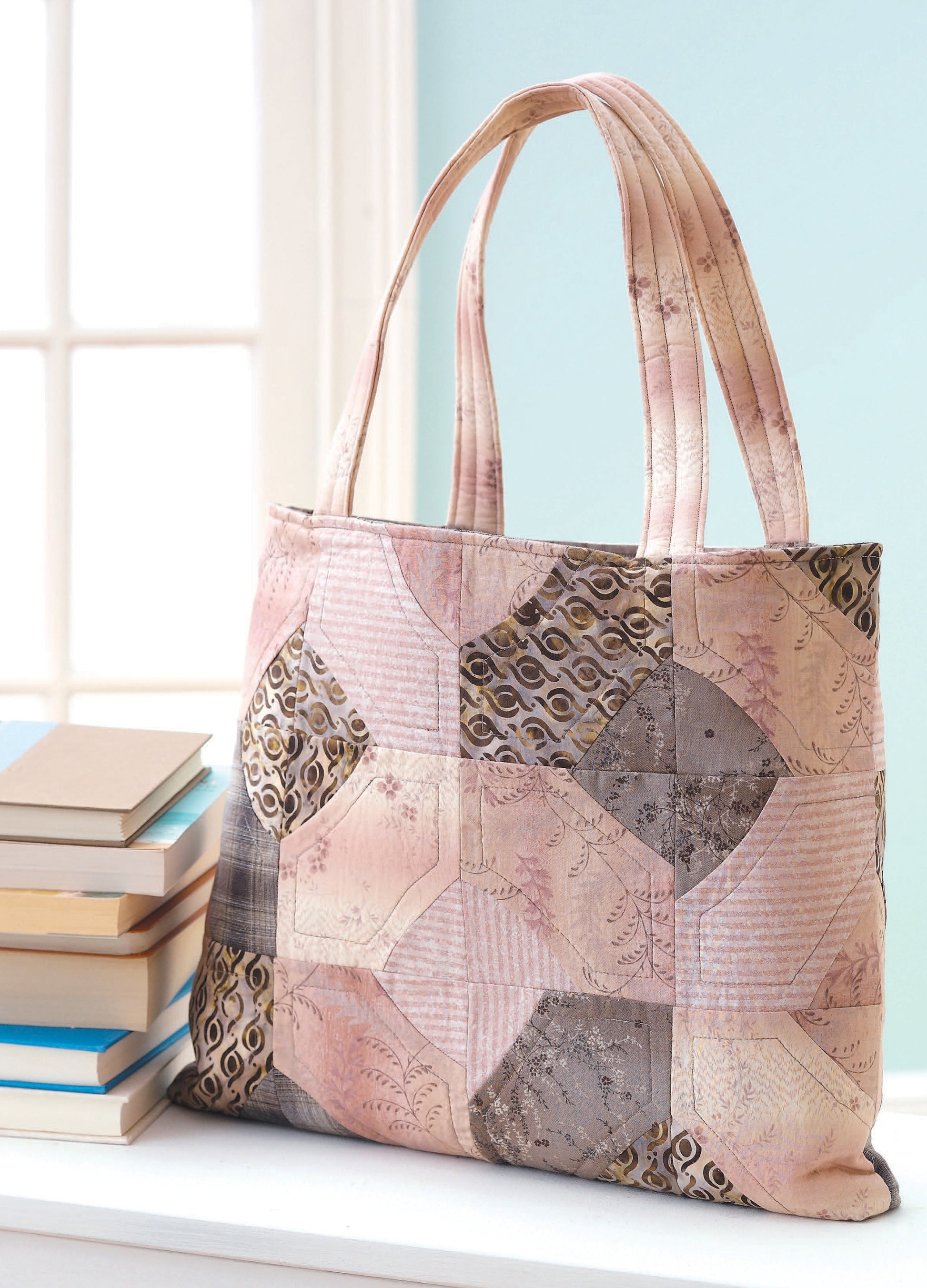The Fat Quarter Bags by Leisure Arts, featuring pastel pink, beige, and gray geometric patchwork, sits on a white surface near a stack of books by a window bathed in soft natural light—a perfect pick for quilted bag fans.