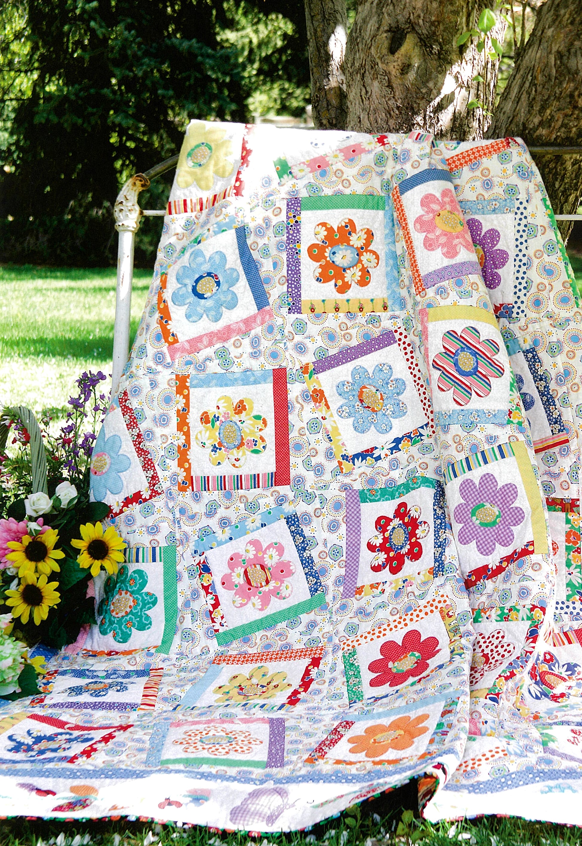 The "Fresh & Fun Applique—12 Projects" by Leisure Arts features a vibrant patchwork quilt with floral appliqué, displayed outdoors on a garden bench beside yellow and purple flowers, bathed in sunlight for year-round charm.