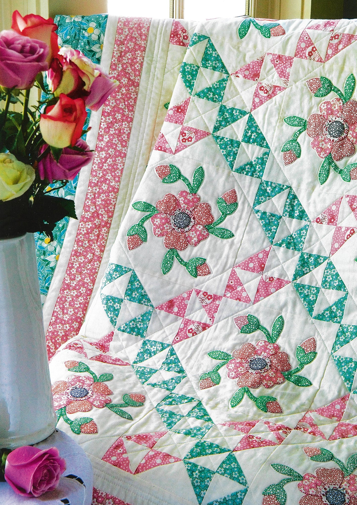 The Leisure Arts “Fresh & Fun Applique—12 Projects” quilt features pink, teal, and white geometric patterns with applique flowers, draped over a chair beside roses, brightening your room with year-round beauty.