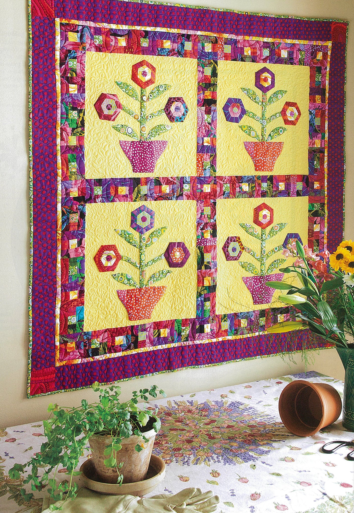 Leisure Arts' Fresh & Fun Applique—12 Projects displays a vibrant patchwork quilt with four flower basket applique designs and hexagon flowers on yellow, bordered by colorful prints, hanging above a table with potted plants and a tipped clay pot.