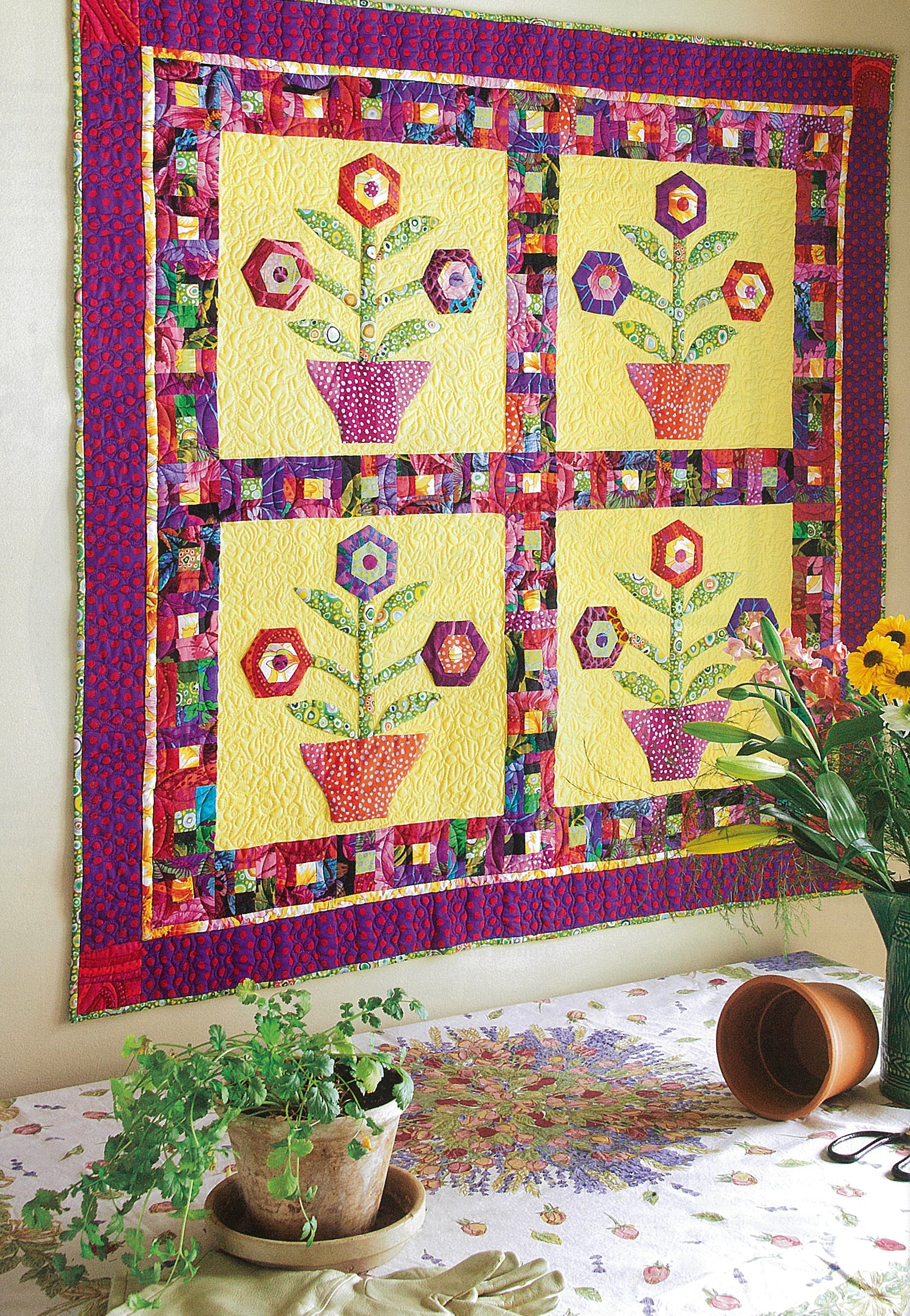 Leisure Arts' Fresh & Fun Applique—12 Projects displays a vibrant patchwork quilt with four flower basket applique designs and hexagon flowers on yellow, bordered by colorful prints, hanging above a table with potted plants and a tipped clay pot.