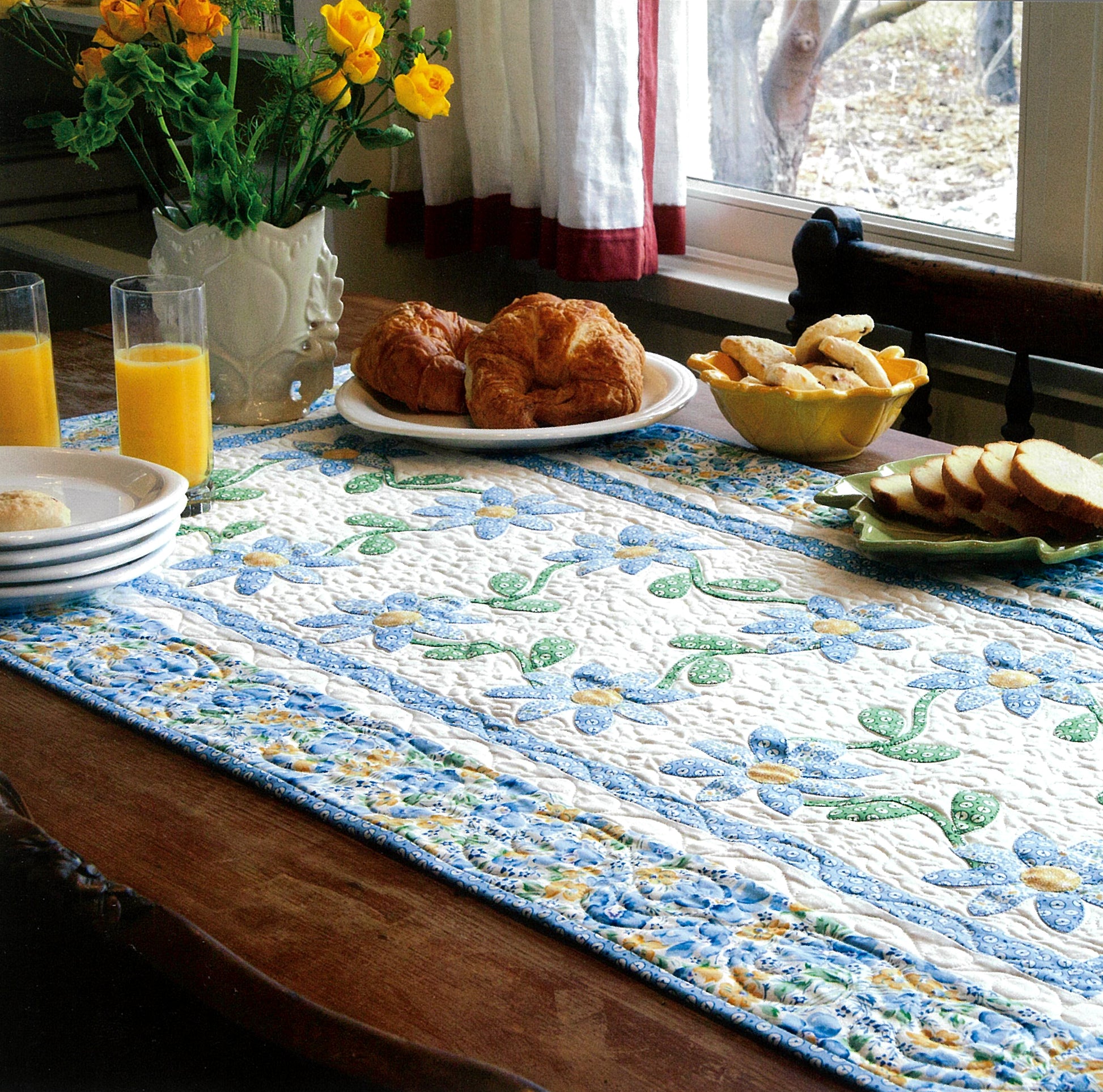 A breakfast table adorned with croissants, juice, and fruit on a quilted runner made from Fresh & Fun Applique—12 Projects by Leisure Arts adds year-round charm near a sunny window with cheerful yellow flowers.