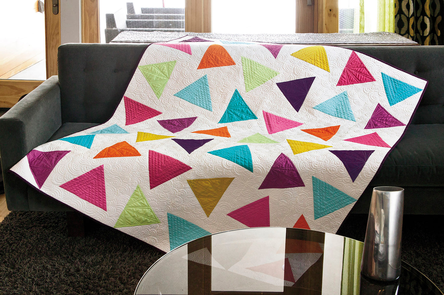 The "Simple Beginner Quilts Digital Download" by Leisure Arts features bright geometric triangles, perfect for adventurous beginners and shown draped over a dark gray sofa in a modern living room with a glass coffee table.