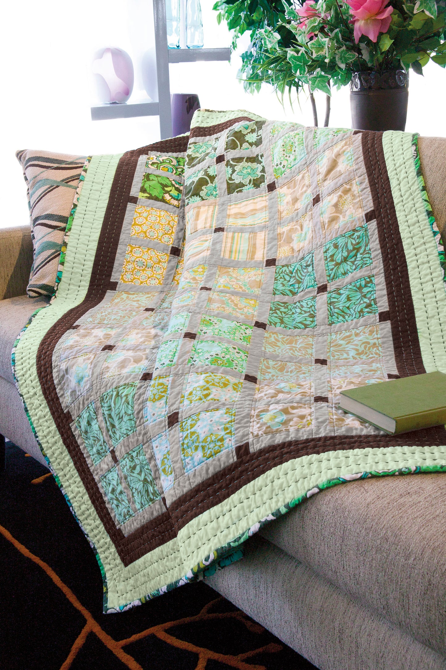A beige couch with striped pillows features a green, yellow, and brown patchwork quilt. The scene highlights Leisure Arts’ Simple Beginner Quilts Digital Download—perfect for adventurous beginners. A closed green book rests nearby.