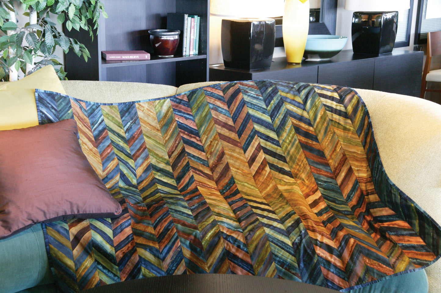The Leisure Arts Simple Beginner Quilts Digital Download features a chevron-patterned earth-tone quilt draped over a yellow couch, inspiring exciting projects for any adventurous beginner in this cozy, modern living room.