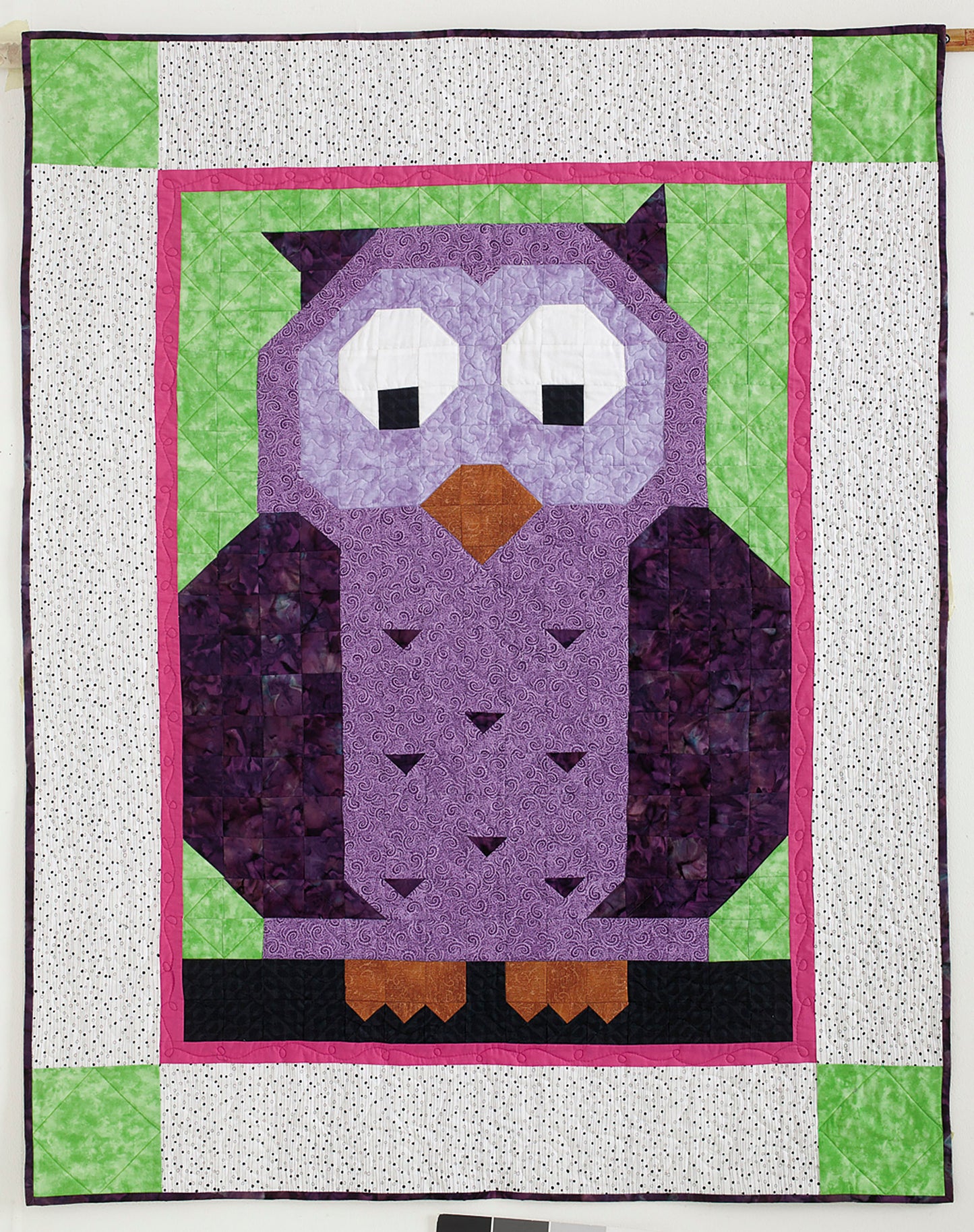 The Patch Pals Collection—Best of Quiltmaker! by Leisure Arts features a handmade quilt with a large purple owl, black and white eyes, orange beak and feet on green, framed by pink and white—ideal for little quilts or beginner quilting projects.