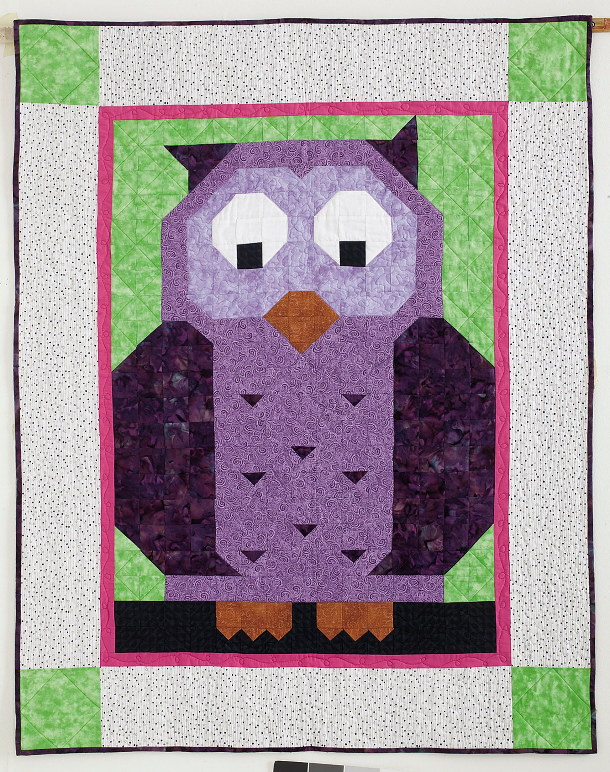 The Patch Pals Collection—Best of Quiltmaker! by Leisure Arts features a handmade quilt with a large purple owl, black and white eyes, orange beak and feet on green, framed by pink and white—ideal for little quilts or beginner quilting projects.