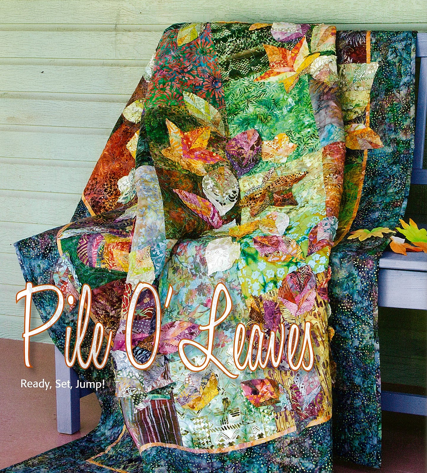A vibrant leaf-themed quilt from “Devoted to Scraps—Best of Quiltmaker” by Leisure Arts is draped over a blue bench against a wooden wall, with decorative “Pile O’ Leaves” and “Ready, Set, Jump!” text at the bottom left.