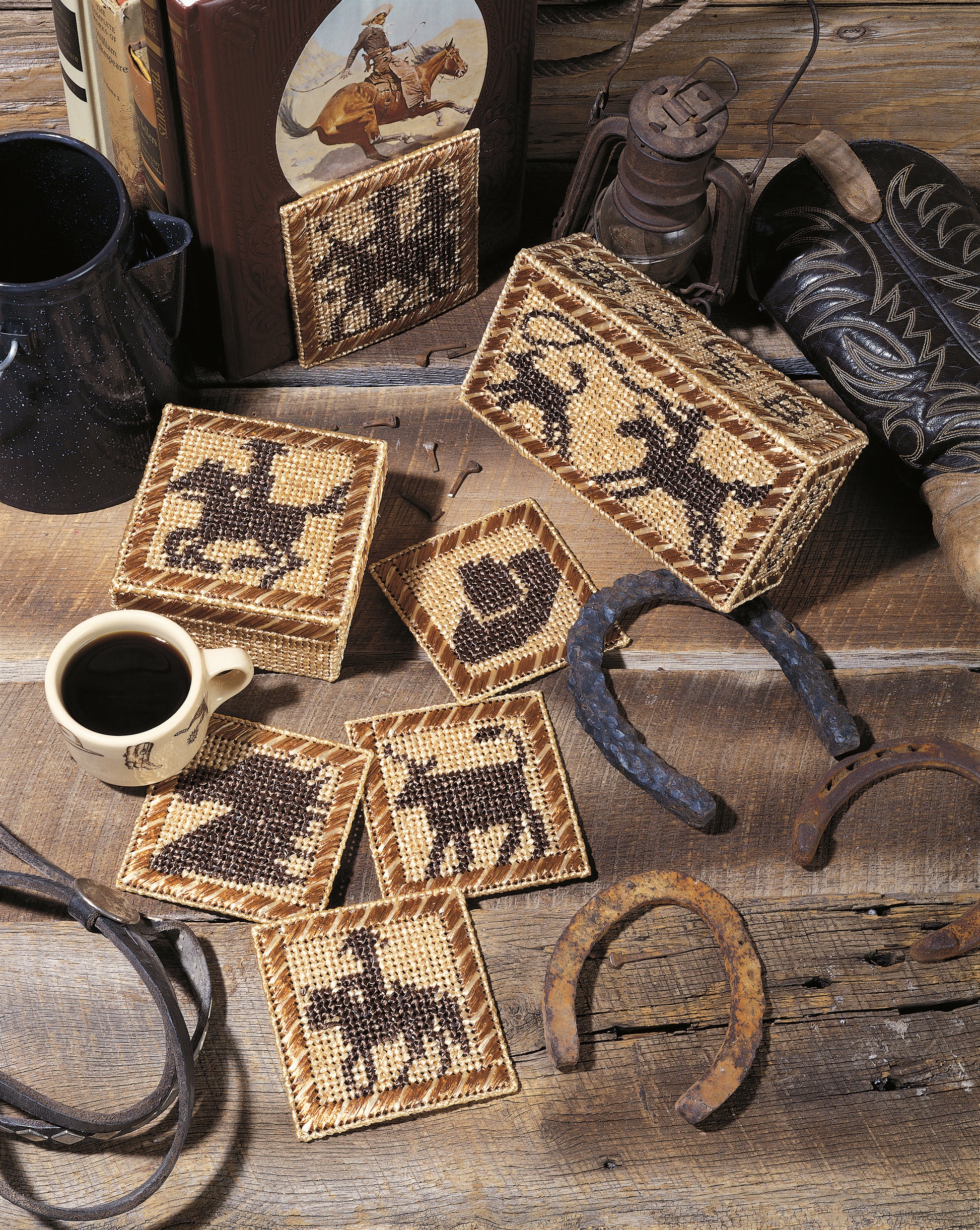 A rustic table displays Leisure Arts’ “Out West—Rustic Designs for the Cowboy in Your Life!” digital download, featuring Wild West motifs like horses, cows, cowboy hats, horseshoes, mugs, lanterns, and rodeo silhouettes.