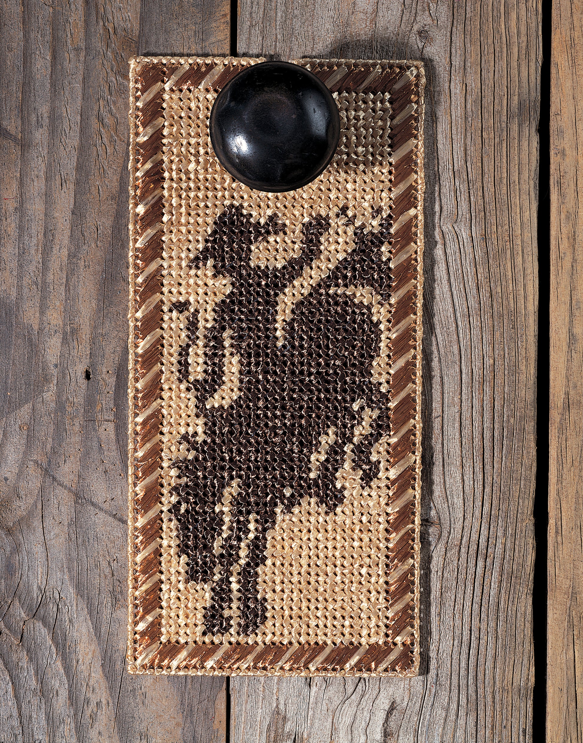 The "Out West—Rustic Designs for the Cowboy in Your Life!" digital download by Leisure Arts features a woven rectangular coaster with a dark cowboy and bucking horse silhouette, zigzag border, and Wild West charm—a small black bowl atop completes the look.