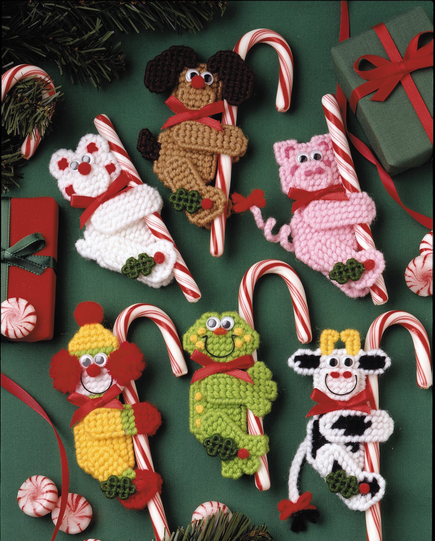 Candy Cane Climbers by Leisure Arts features six crocheted animal ornaments—dog, bear, pig, clown, frog, and cow—each with a candy cane and red ribbon. Displayed with gifts and pine branches, they’re charming Christmas crafts.