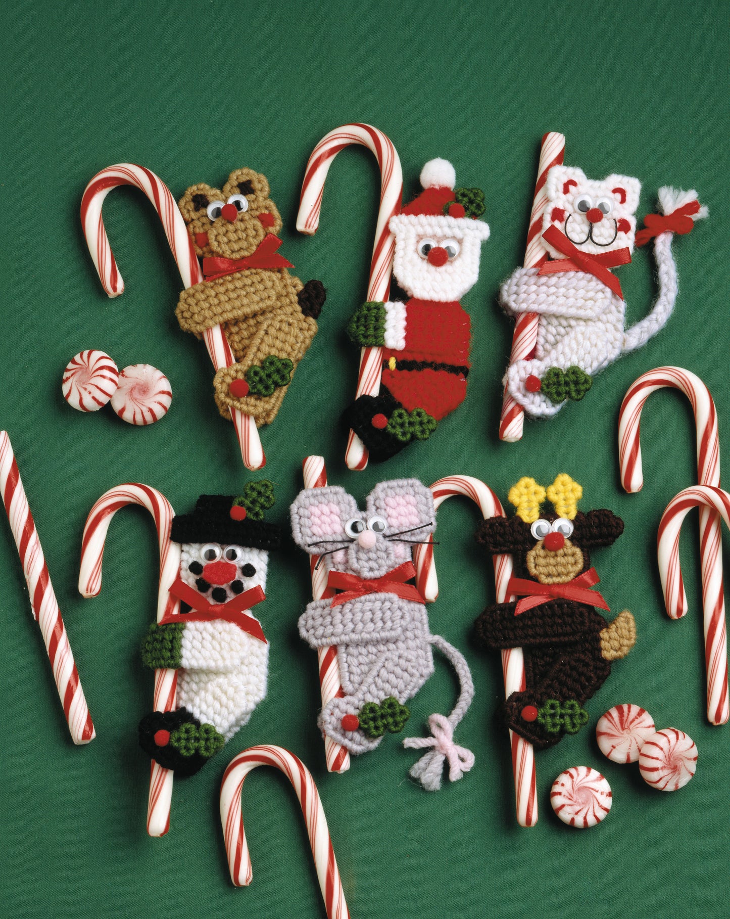 Candy Cane Climbers by Leisure Arts features six crocheted Christmas characters (bear, Santa, cat, snowman, mouse, and reindeer) attached to candy canes—festive ornaments perfect for holiday crafts—shown on a green surface with extra treats.