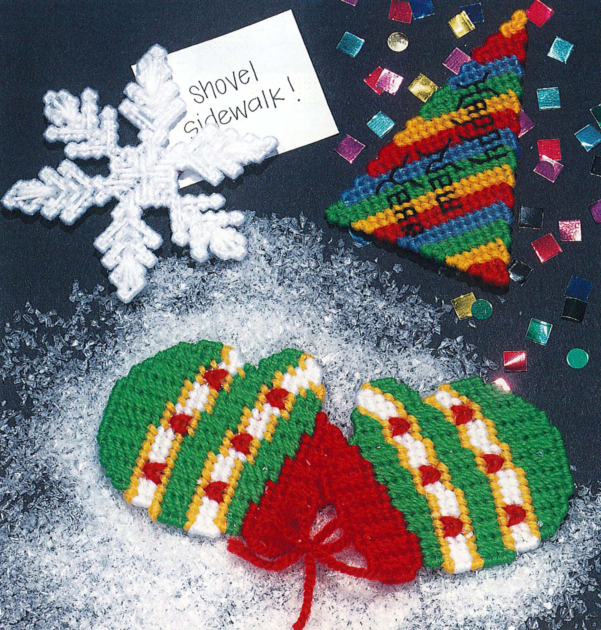 Vibrant knitted Christmas ornaments—a snowflake, striped tree, and green mittens—sit on snow with confetti and a "Shovel sidewalk!" note, perfect for Leisure Arts’ Magnets Thru Out the Year seasonal plastic canvas projects.