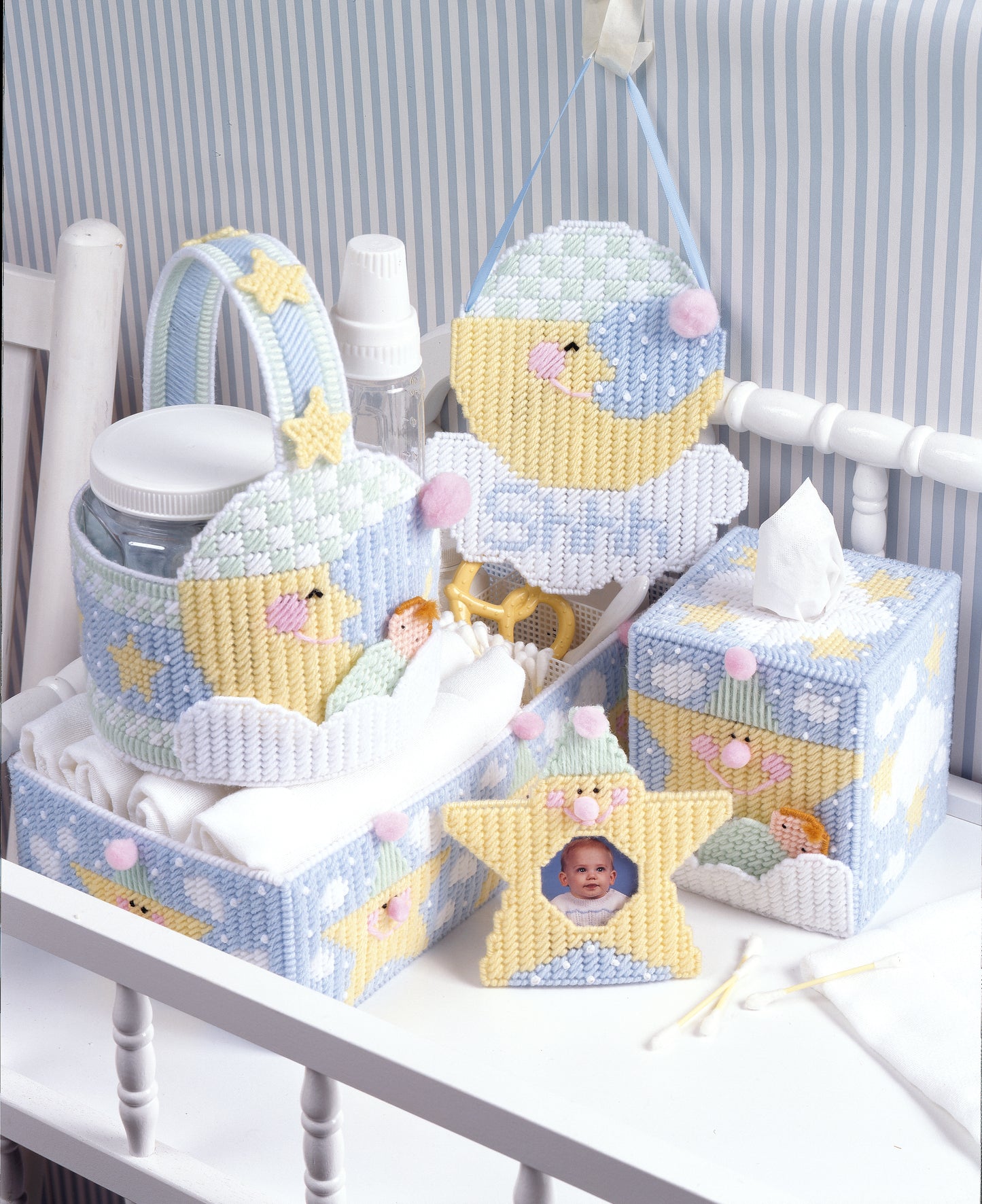 A Star Is Born by Leisure Arts offers pastel blue, yellow, and white plastic canvas nursery accessories—tissue box, containers, and moon and star cloud decor—perfectly displayed on a white crib rail for your tot's picture perfect nursery.