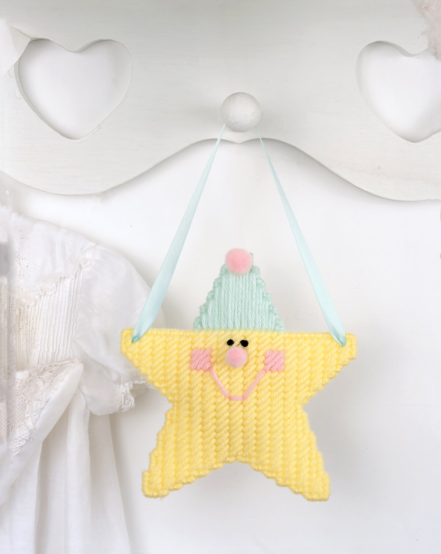 A Star Is Born by Leisure Arts—a soft yellow star-shaped ornament with a smiling face, pink cheeks, and blue hat hangs on a ribbon amid heart cutouts for perfect nursery décor and charming decorative accents.