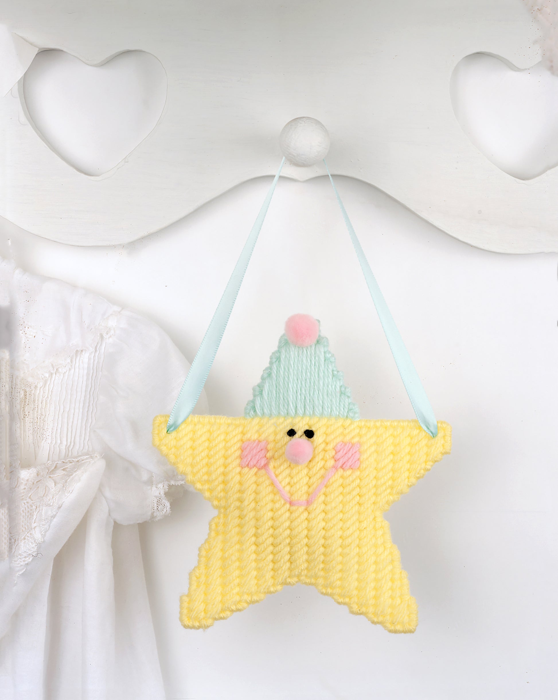 A Star Is Born by Leisure Arts—a soft yellow star-shaped ornament with a smiling face, pink cheeks, and blue hat hangs on a ribbon amid heart cutouts for perfect nursery décor and charming decorative accents.