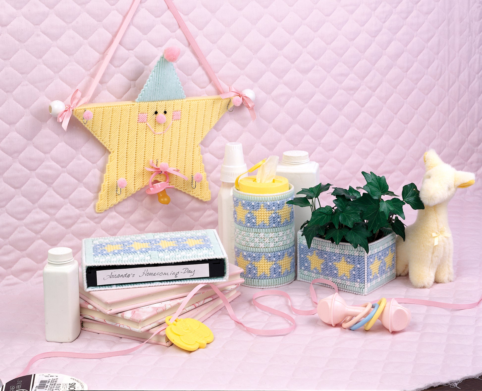 Create a picture-perfect nursery with “A Star Is Born” by Leisure Arts. Shown on a pink quilted background: yellow star decor, baby bottles, plush giraffe, pacifier, rattle, photo albums, decorative accents & a small plant.
