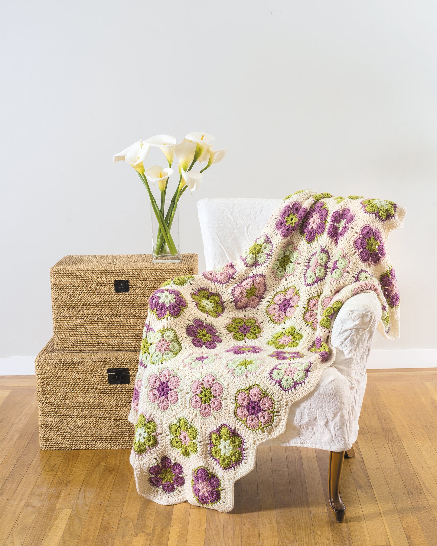 A white armchair with a crochet blanket inspires your next project using Leisure Arts' "Learn to Crochet African Flower Motifs" digital download. Nearby, two wicker boxes and calla lilies on wood flooring complete the scene against a plain wall.