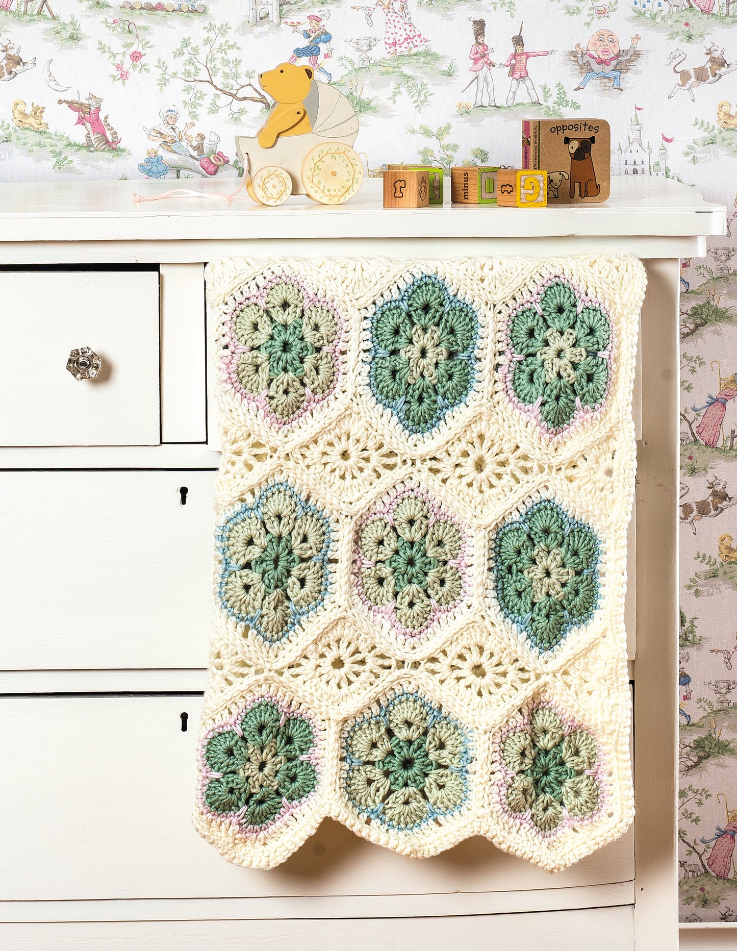 A crocheted blanket made with the “Learn to Crochet African Flower Motifs” digital download by Leisure Arts, featuring green, blue, and purple flowers in worsted yarn, hangs from a cream dresser topped with animal toys and alphabet blocks; vintage wallpaper behind.