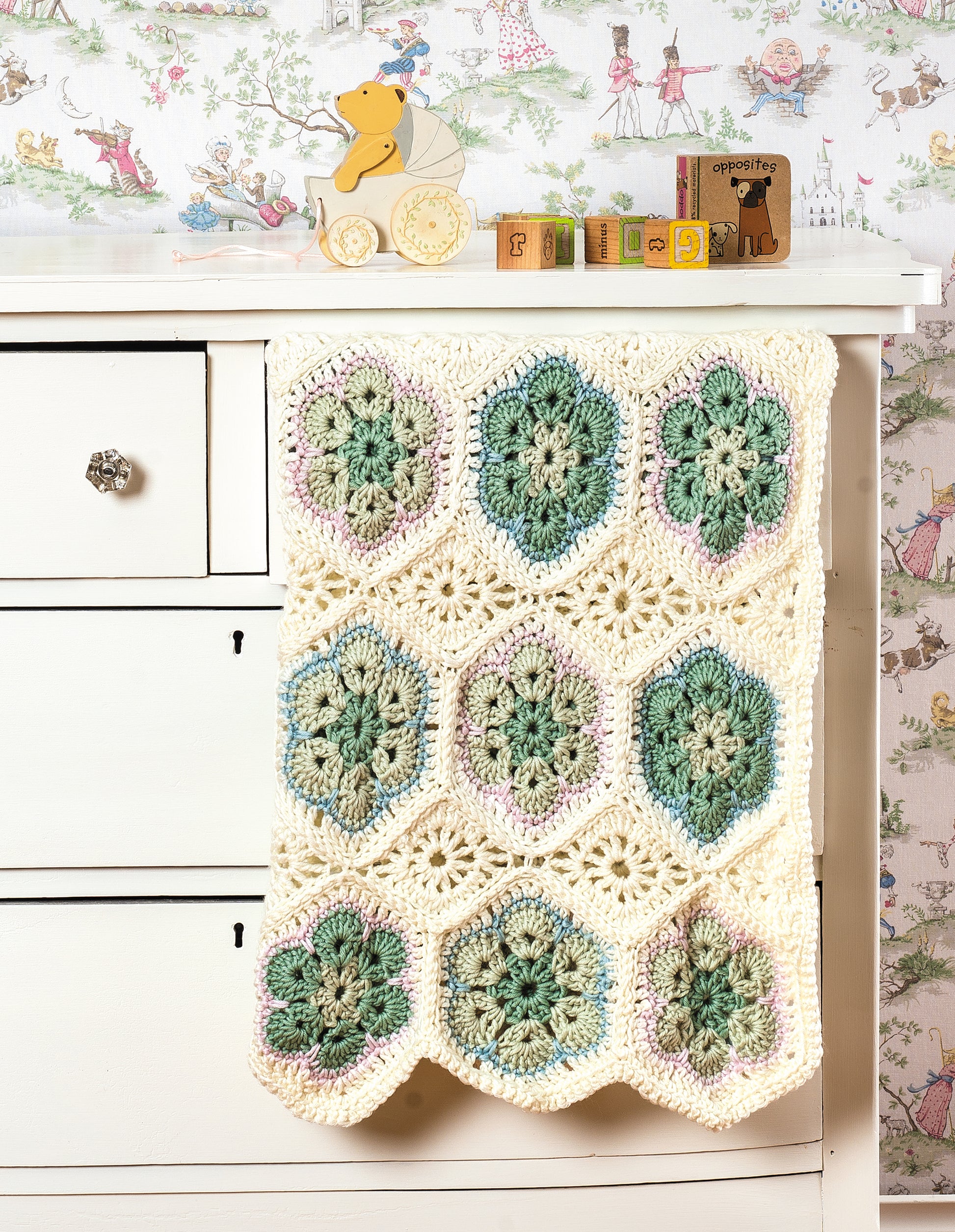 A crocheted blanket made with the “Learn to Crochet African Flower Motifs” digital download by Leisure Arts, featuring green, blue, and purple flowers in worsted yarn, hangs from a cream dresser topped with animal toys and alphabet blocks; vintage wallpaper behind.