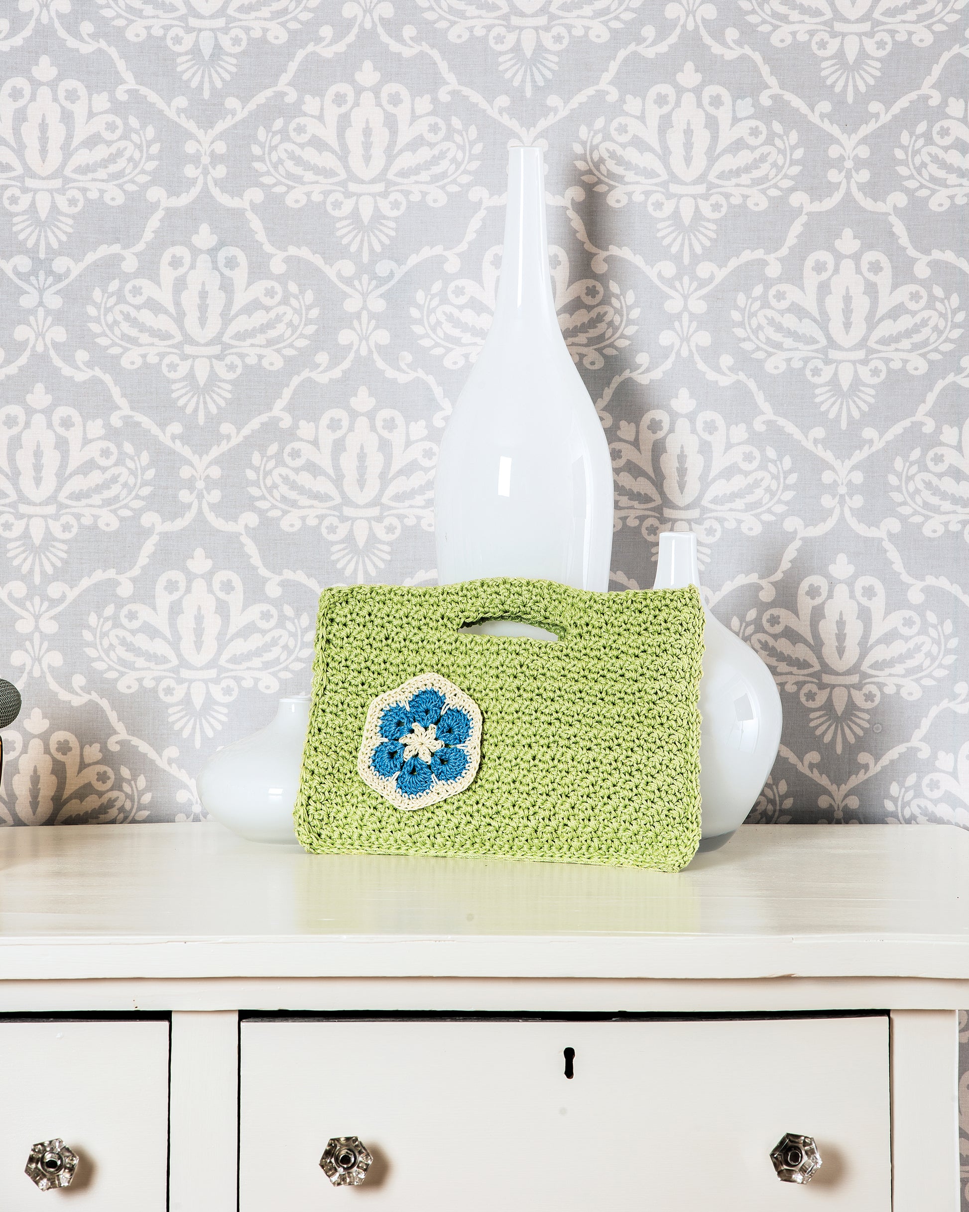 The "Learn to Crochet African Flower Motifs" digital download by Leisure Arts showcases a light green clutch with blue and white flower motifs—perfect inspiration for your next crochet project. Displayed elegantly on a white cabinet with vases and gray wallpaper.