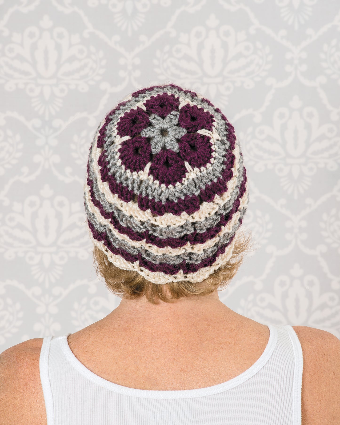 A person in a white tank top faces away, showcasing a hat featuring maroon, cream, and gray motifs made using the "Learn to Crochet African Flower Motifs Digital Download" by Leisure Arts against a light gray decorative wallpaper.