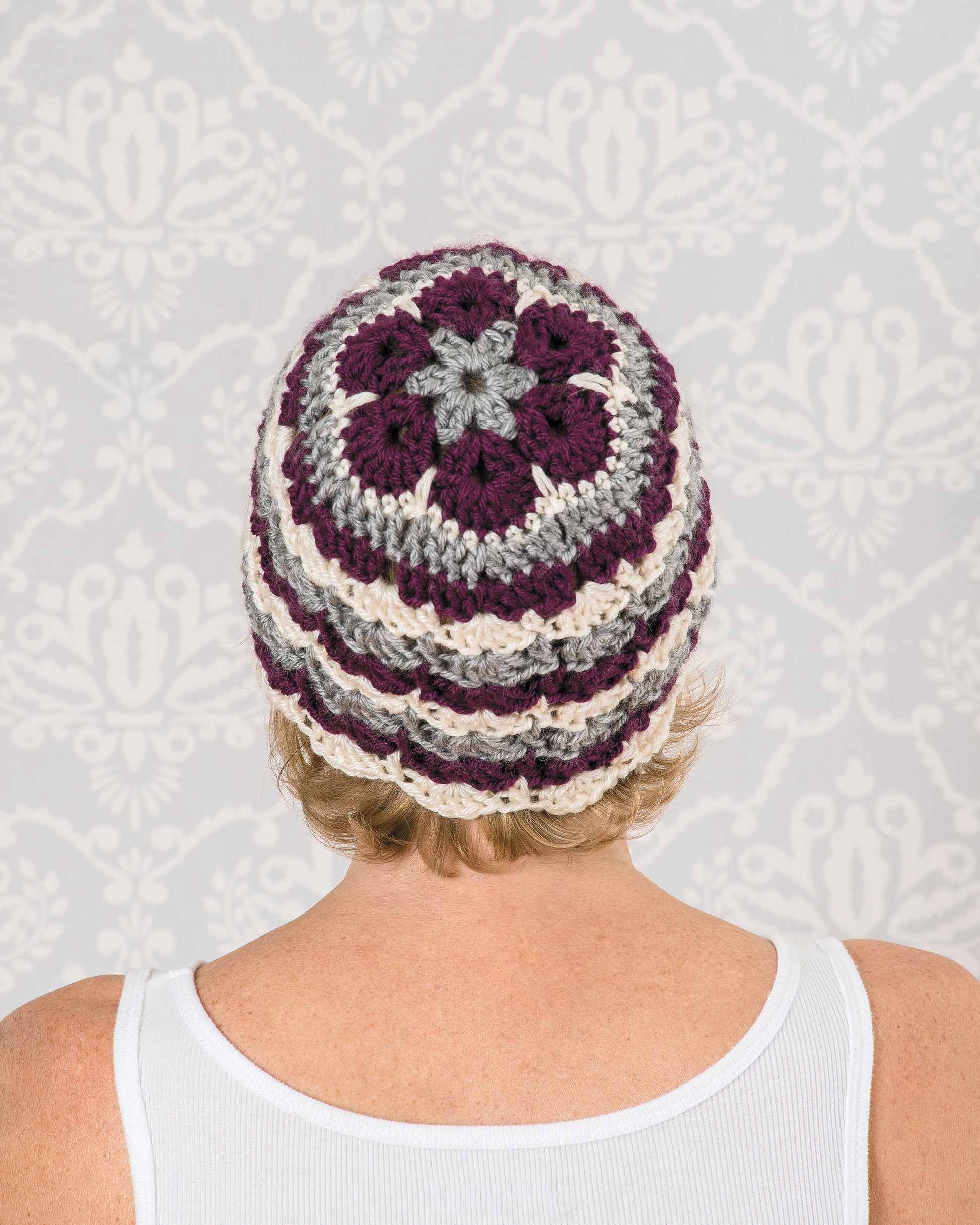 A person in a white tank top faces away, showcasing a hat featuring maroon, cream, and gray motifs made using the "Learn to Crochet African Flower Motifs Digital Download" by Leisure Arts against a light gray decorative wallpaper.