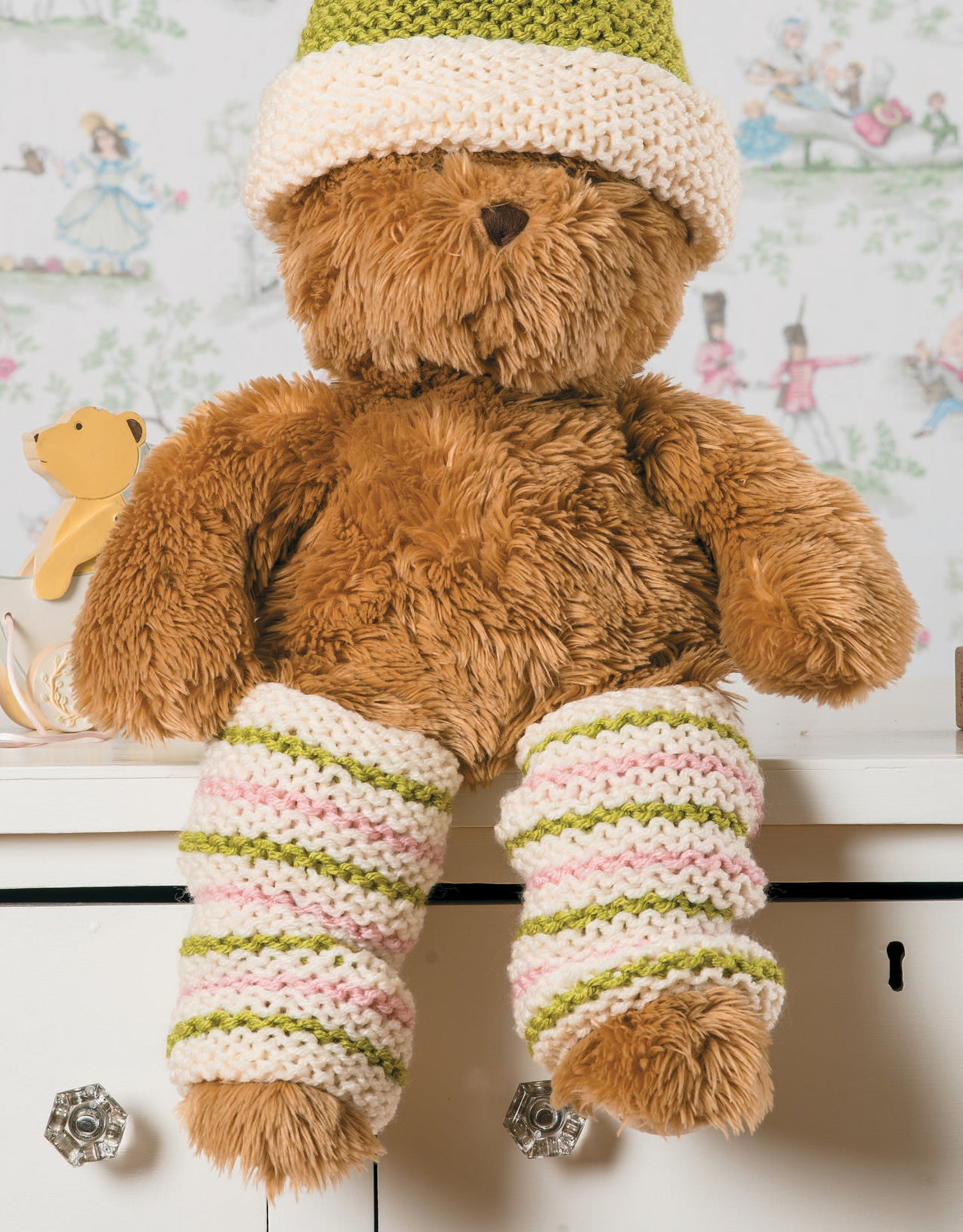 A brown teddy bear in green and white garter stitch knit accessories from Garter Stitch for Baby by Leisure Arts sits on a white dresser, with a yellow toy dog and illustrated wallpaper in the background.