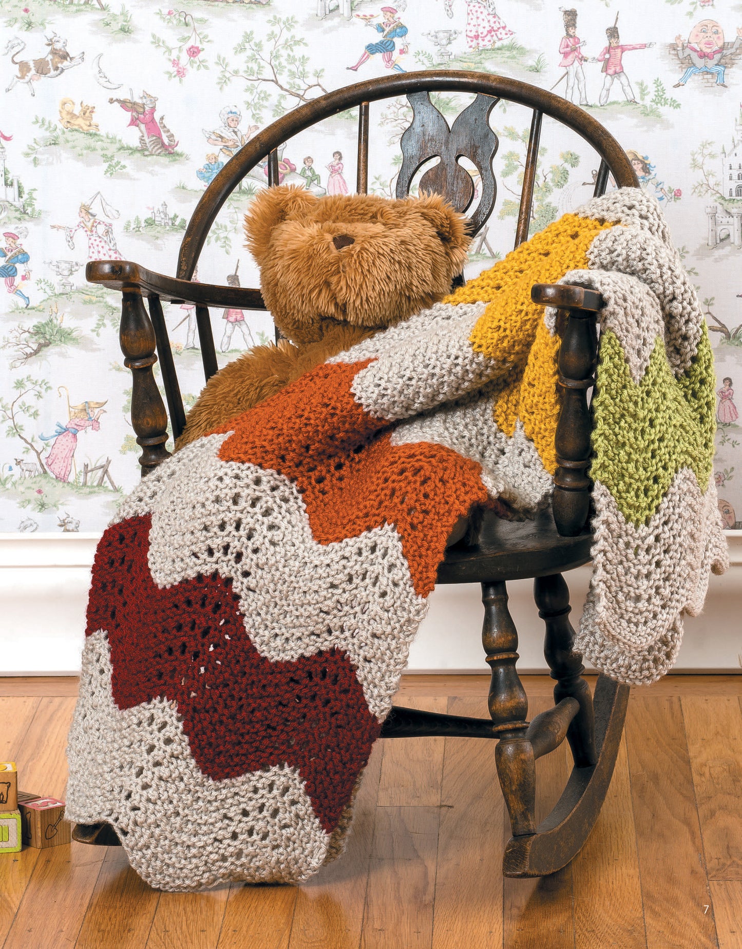 A brown teddy bear sits on a wooden rocking chair draped with a zigzag-patterned knit blanket in beige, rust, yellow, and green made using "Garter Stitch for Baby" by Leisure Arts. The background shows vintage-style illustrated wallpaper.