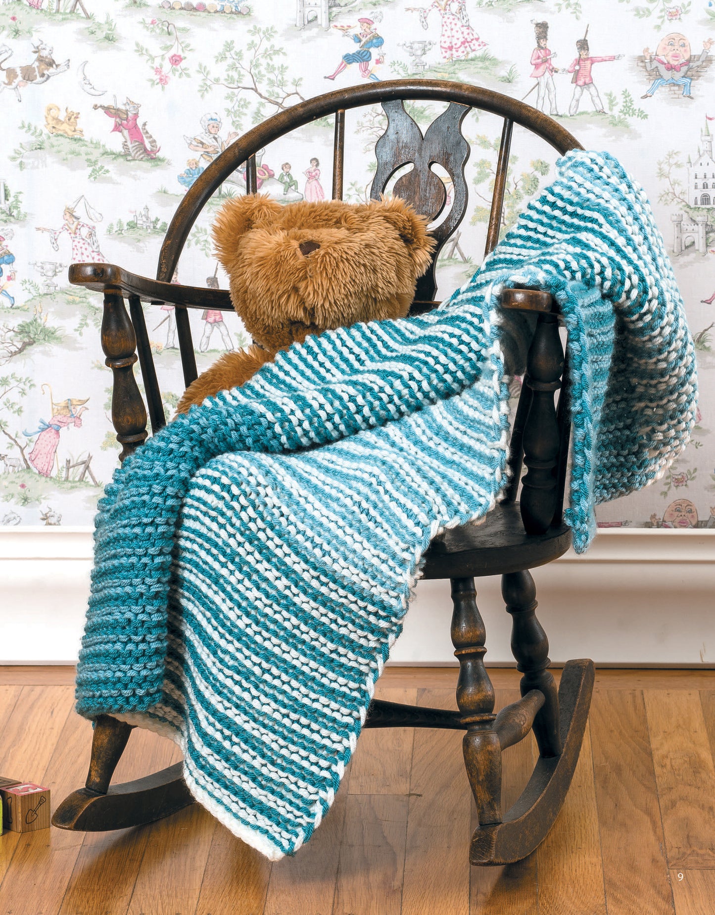 A brown teddy bear sits on a wooden rocking chair draped with a blue and white striped blanket from "Garter Stitch for Baby" by Leisure Arts, set on a wooden floor with whimsical illustrated wallpaper in the background.