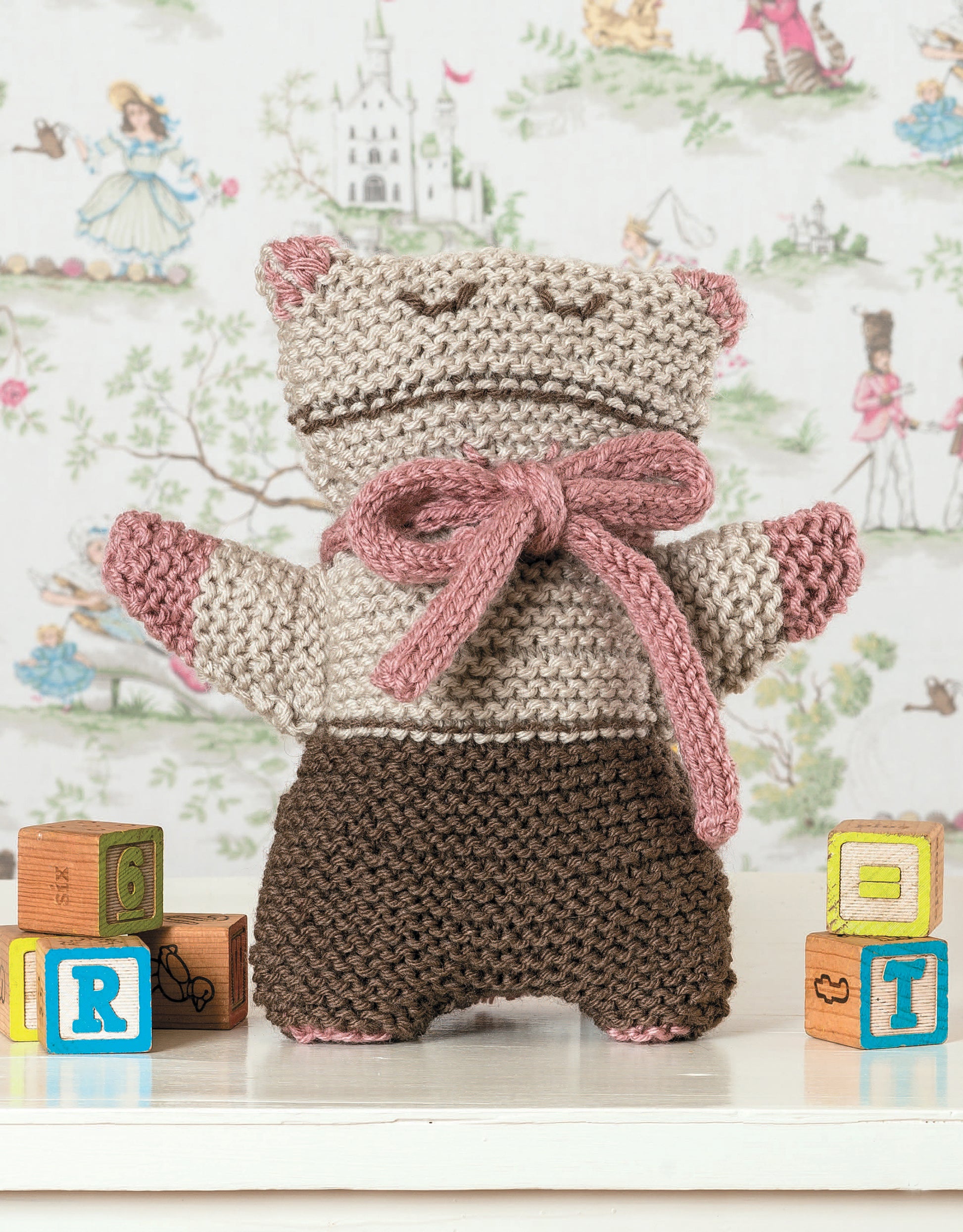 A knitted stuffed animal from "Garter Stitch for Baby" by Leisure Arts stands on a white surface near colorful alphabet blocks, seen from the back with a pink bow and whimsical children's wallpaper in the background.