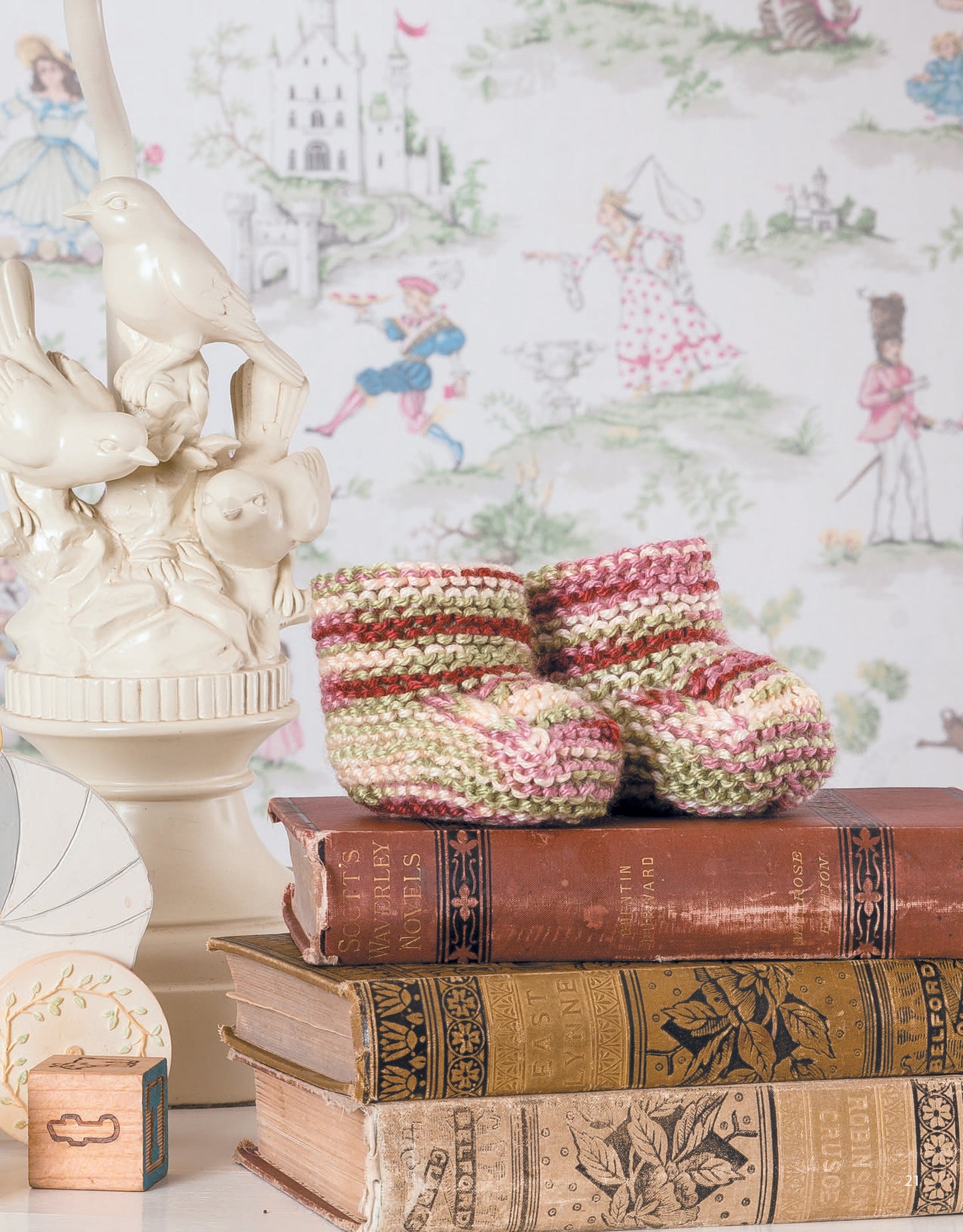 Leisure Arts’ "Garter Stitch for Baby" features pink and green baby booties—a sweet beginner project—displayed on vintage books beside an ornate ceramic lamp, set against whimsical fairytale wallpaper.