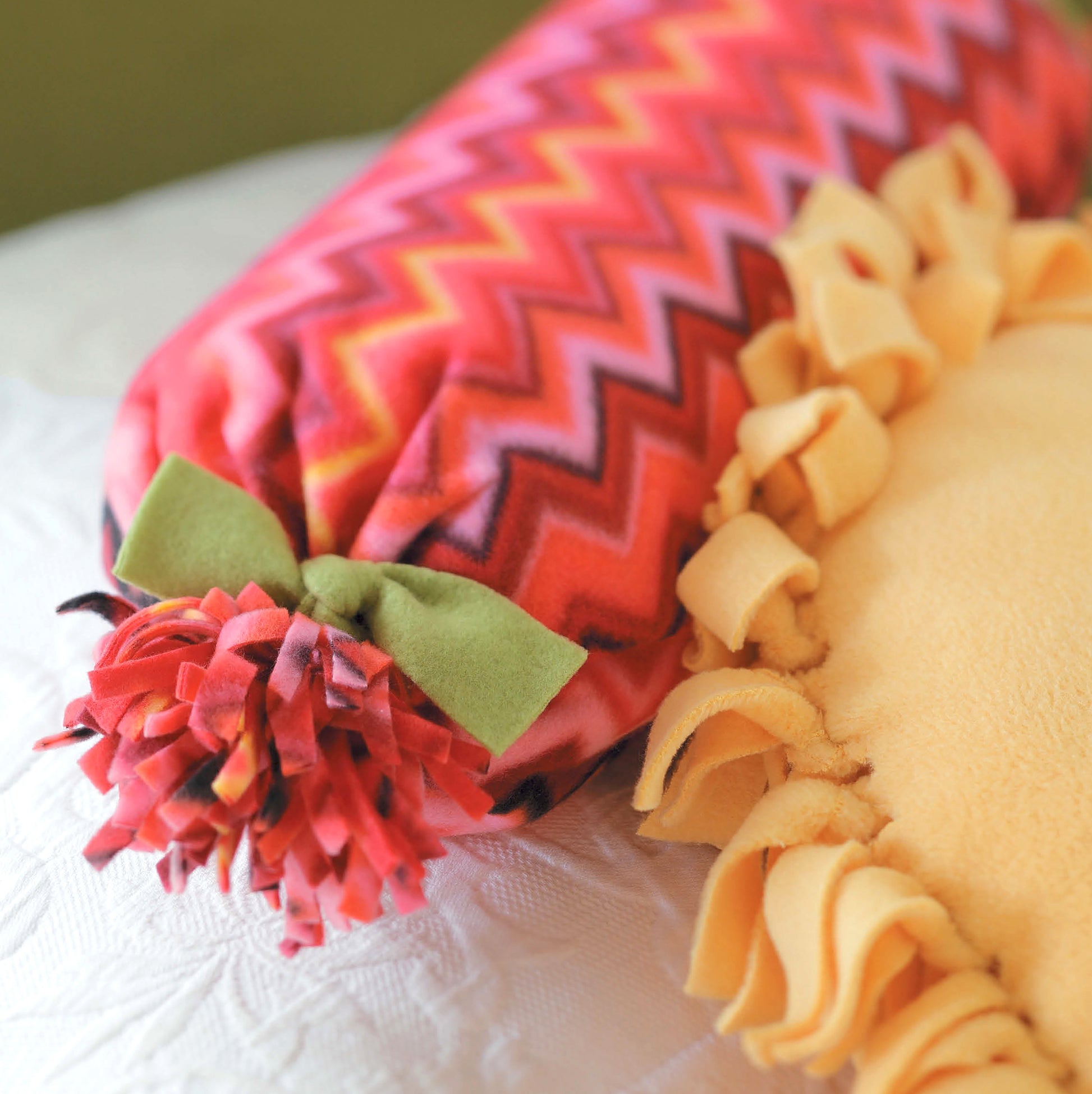 The No-Sew Fleece Throws & Pillows by Leisure Arts showcases a vibrant cylindrical pillow with a zigzag pattern, green bow, and tasseled end beside a yellow knotted-edge fleece pillow—ideal for beginner-friendly home decor crafts.