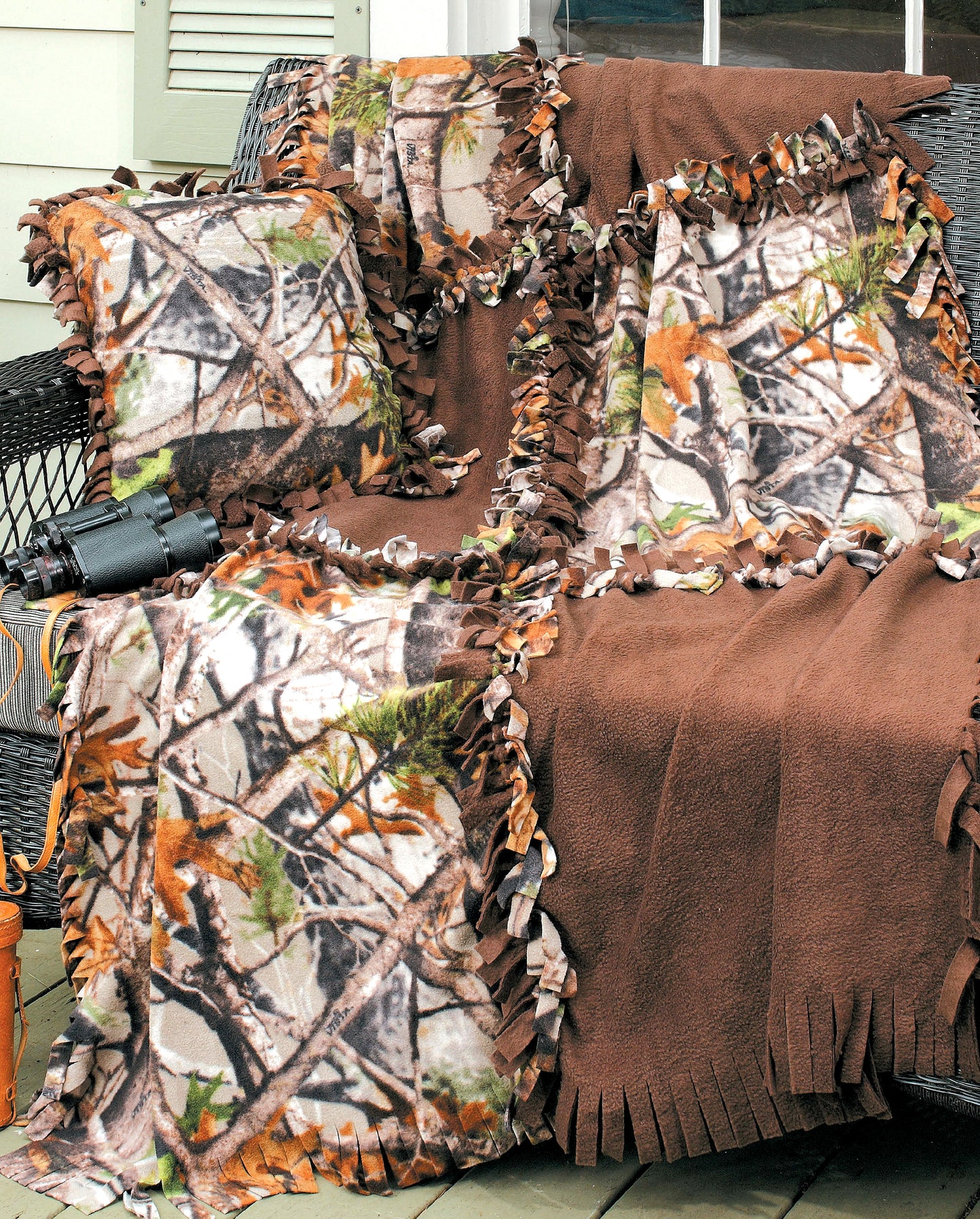 On a wooden deck, Leisure Arts’ No-Sew Fleece Throws & Pillows in brown and camouflage patterns add cozy charm—perfect for beginner fleece crafts—alongside a pair of binoculars and part of a tan bag.