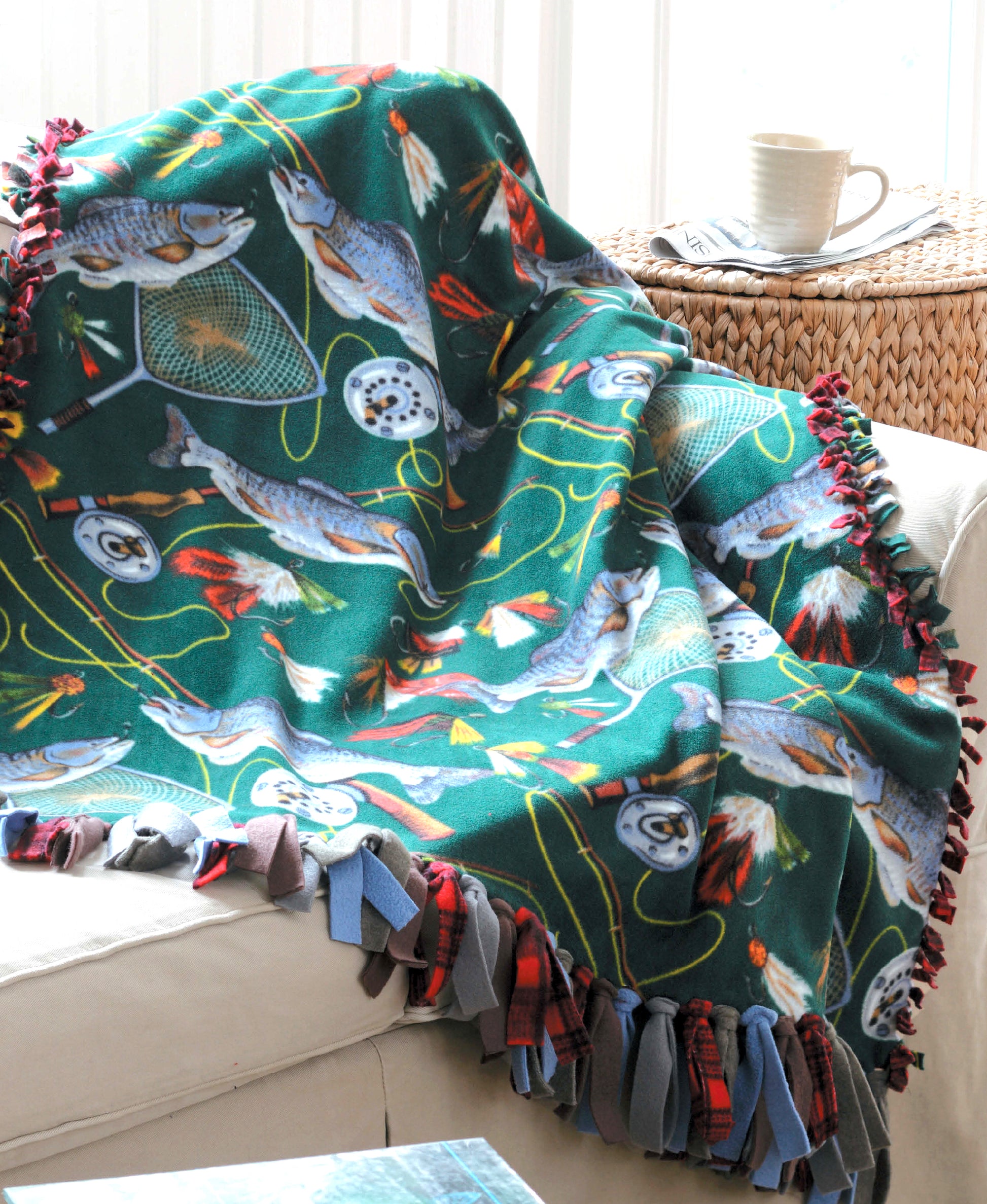 The Leisure Arts No-Sew Fleece Throws & Pillows, with a fishing theme of fish, reels, and lures, brings colorful style to fleece home decor—perfect draped over a light sofa next to a wicker table and white mug.