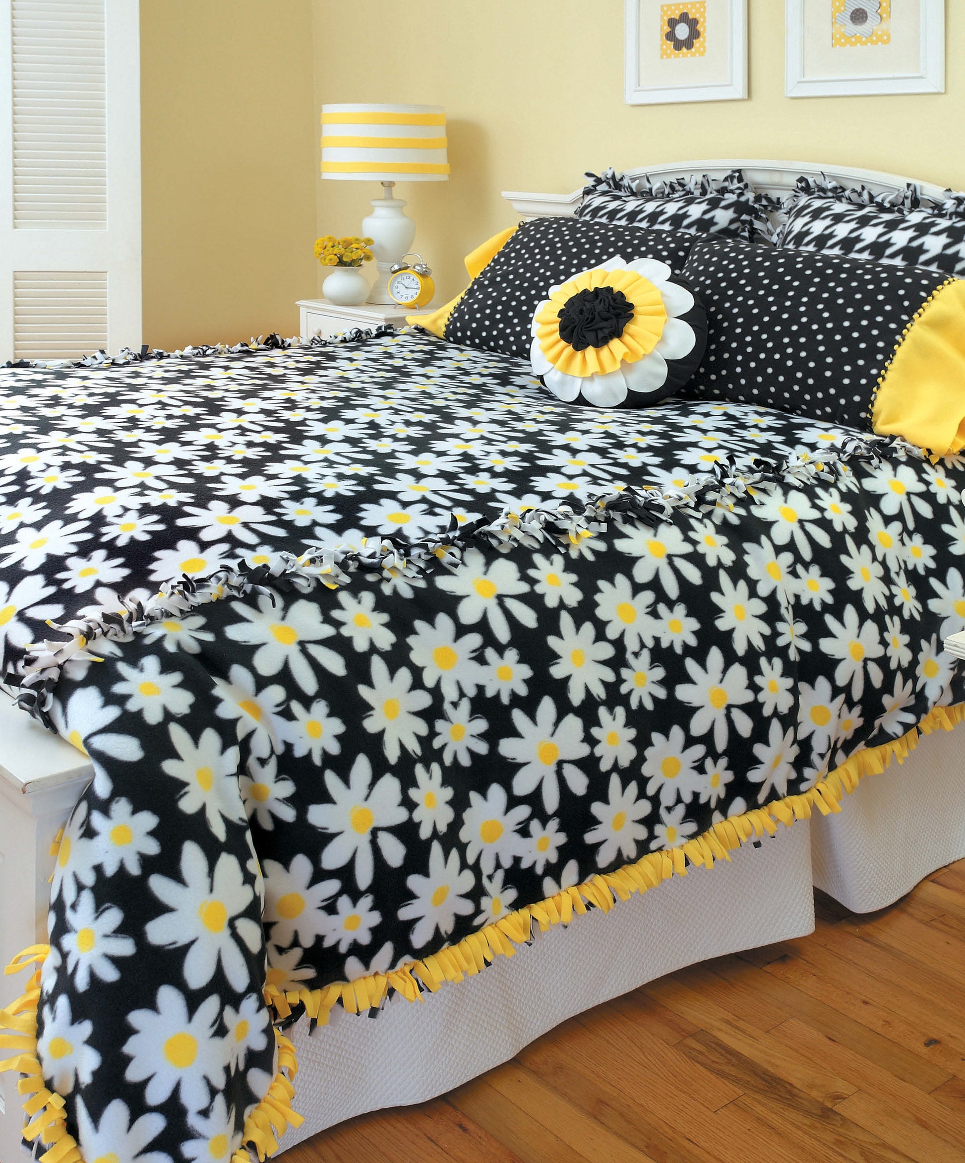 A bed features Leisure Arts' No-Sew Fleece Throws & Pillows—black fleece with white and yellow daisies, matching pillows, and a daisy accent pillow—an inviting, beginner-friendly home decor craft in a yellow, white, and black room.