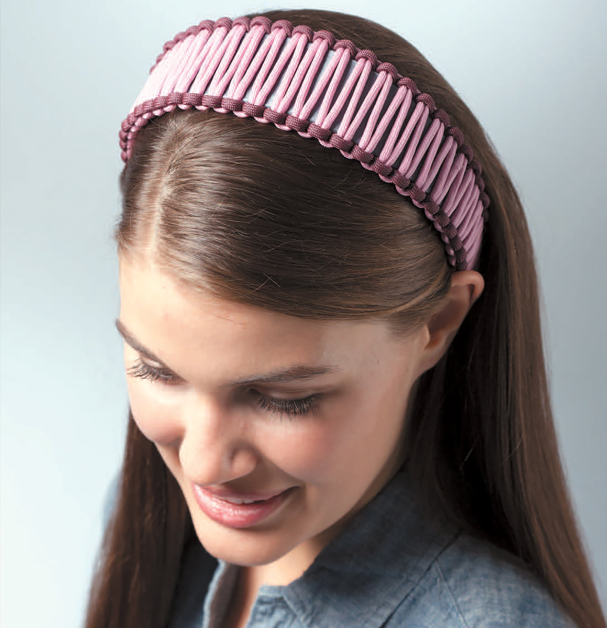 A young woman with straight brown hair wears a wide, pink, woven Paracord Crafts headband by Leisure Arts. She looks down and smiles slightly in a blue shirt against a light background.