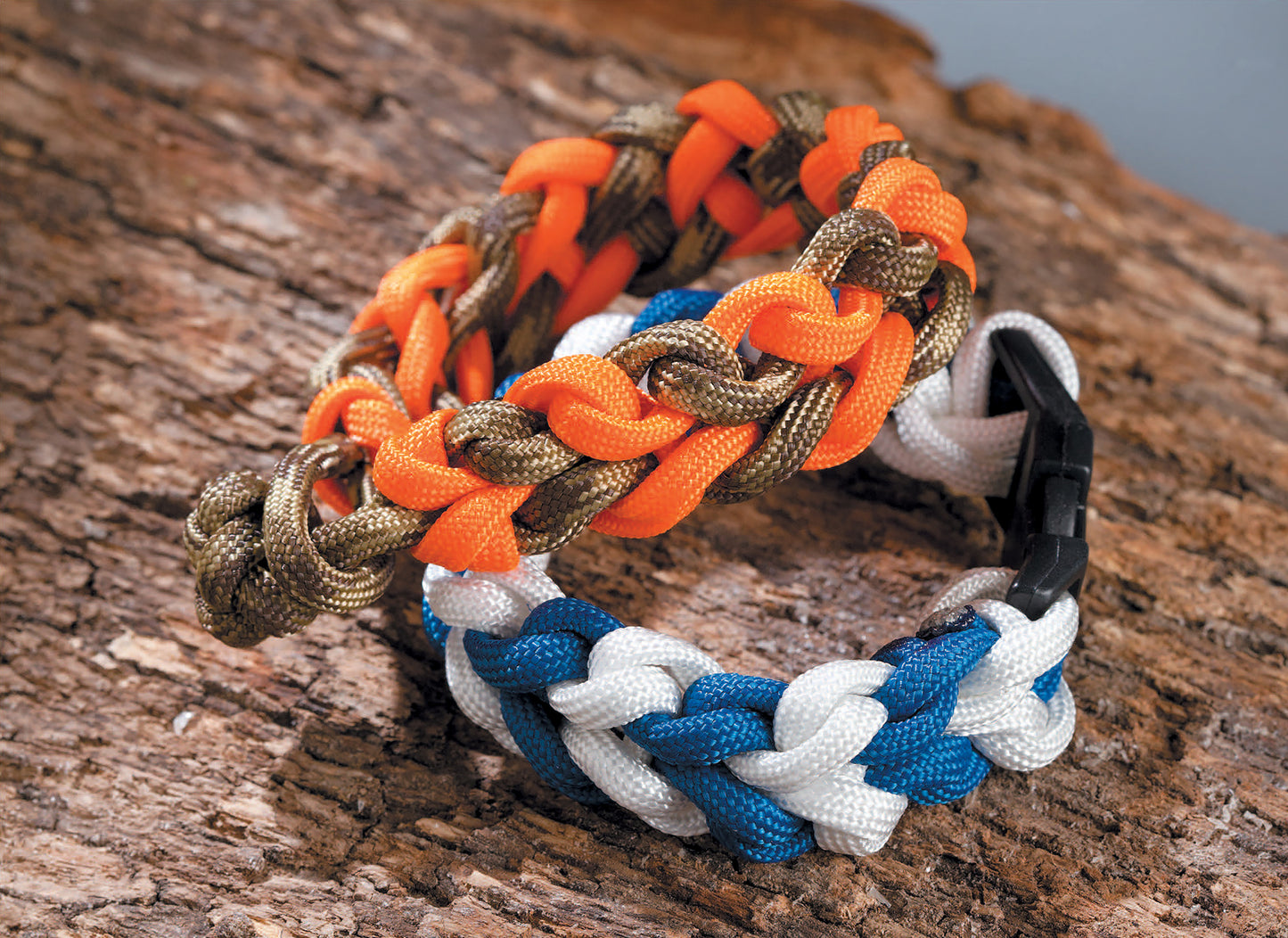 Two woven Paracord Crafts bracelets by Leisure Arts, made with the classic cobra stitch, rest on tree bark—one in orange and brown, the other blue and white with a black buckle. Great for paracord projects or everyday style.